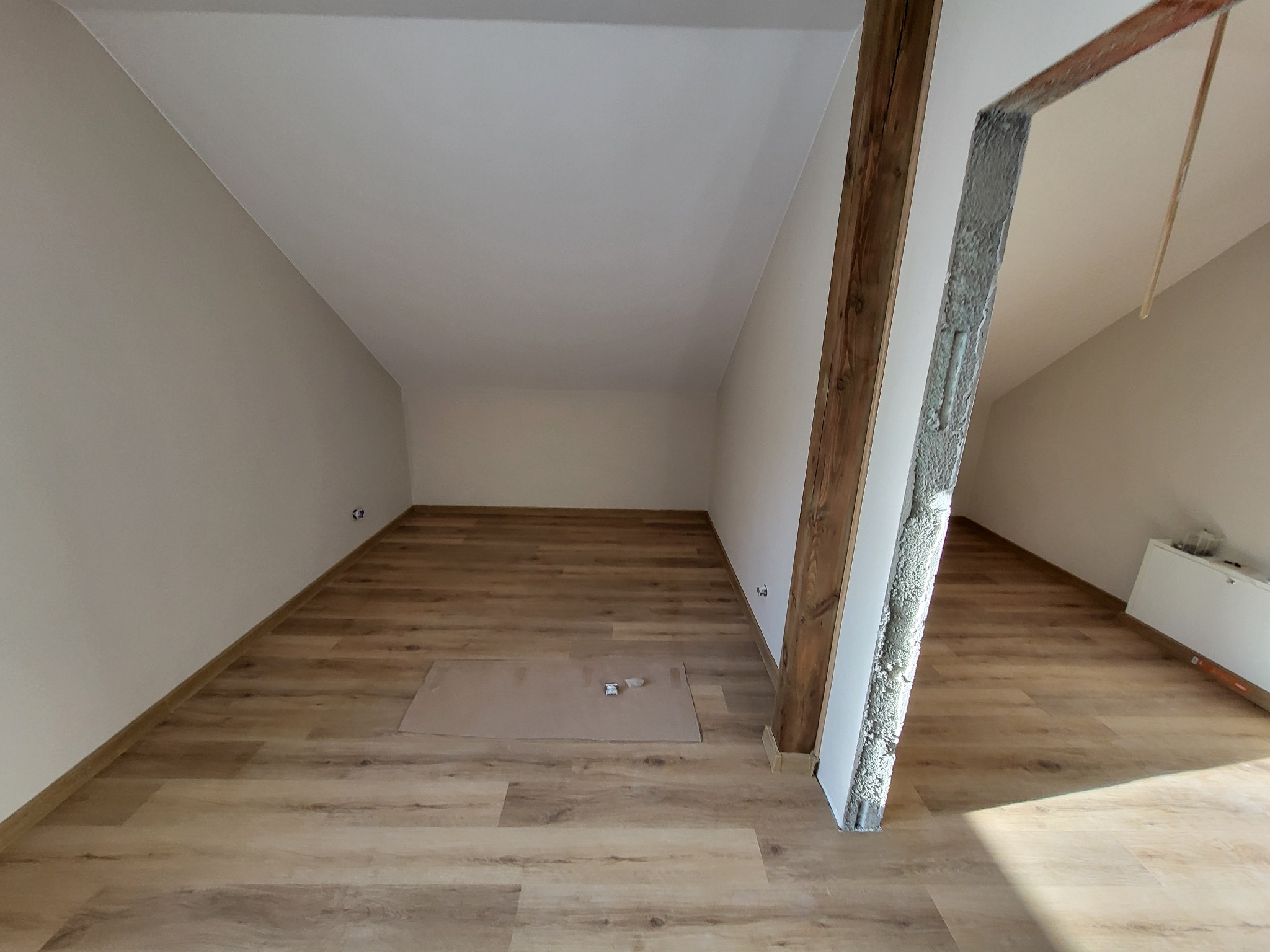 Poddasze po remoncie z nową podłogą z paneli winylowych i świeżo pomalowanymi ścianami, widoczna drewniana belka konstrukcyjna i surowa ościeżnica drzwiowa.