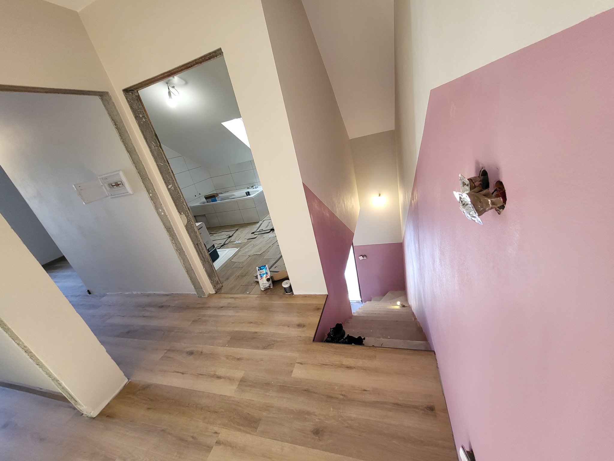 Wnętrze w trakcie remontu: świeżo położone panele winylowe na podłodze, pomalowana na różowo ściana z wystającymi kablami, widok na schody i łazienkę z białymi płytkami w tle.