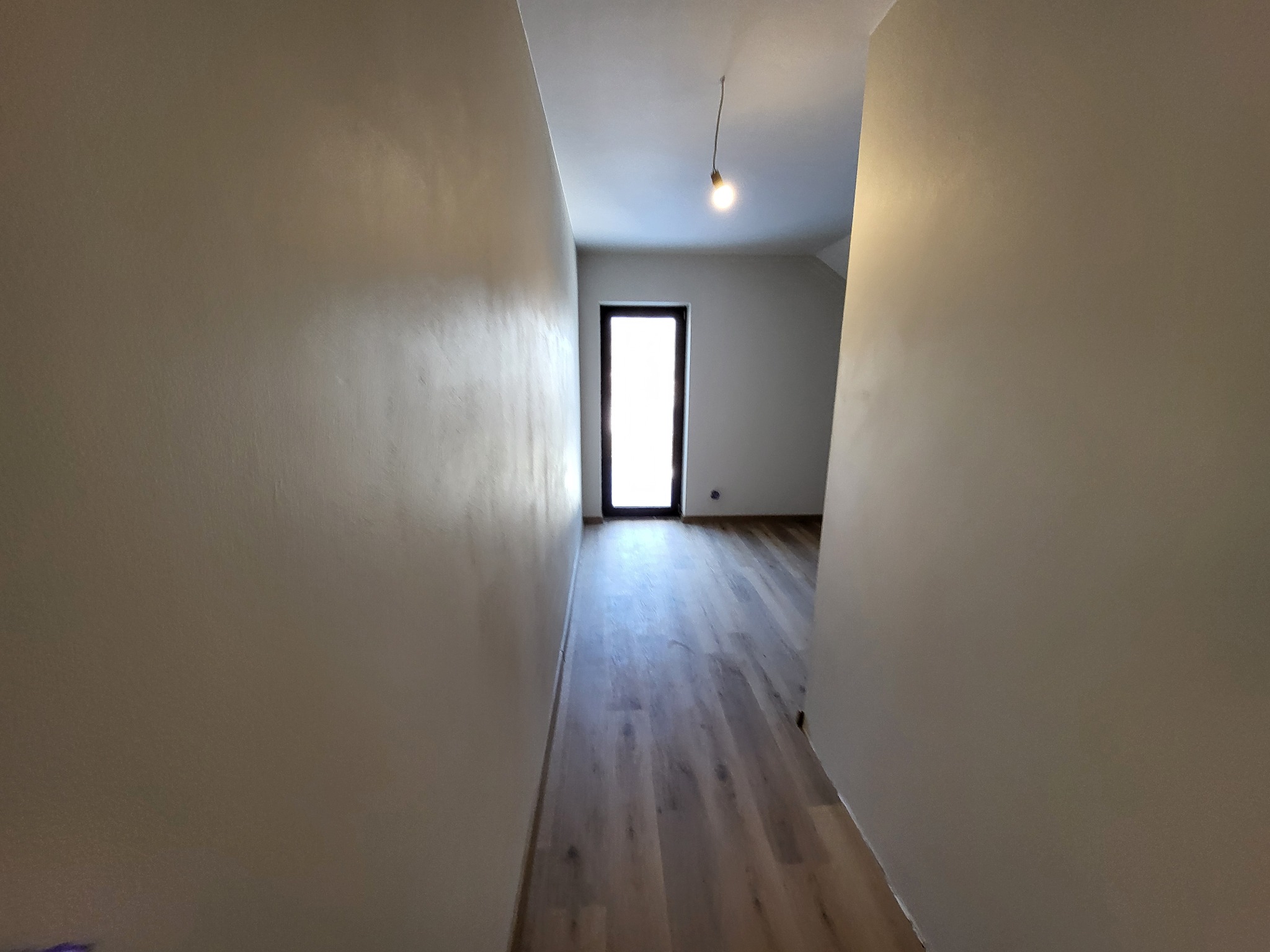 Korytarz po remoncie z położonymi panelami podłogowymi i świeżo wygładzonymi ścianami, oświetlony wiszącą żarówką, widok na drzwi wejściowe.