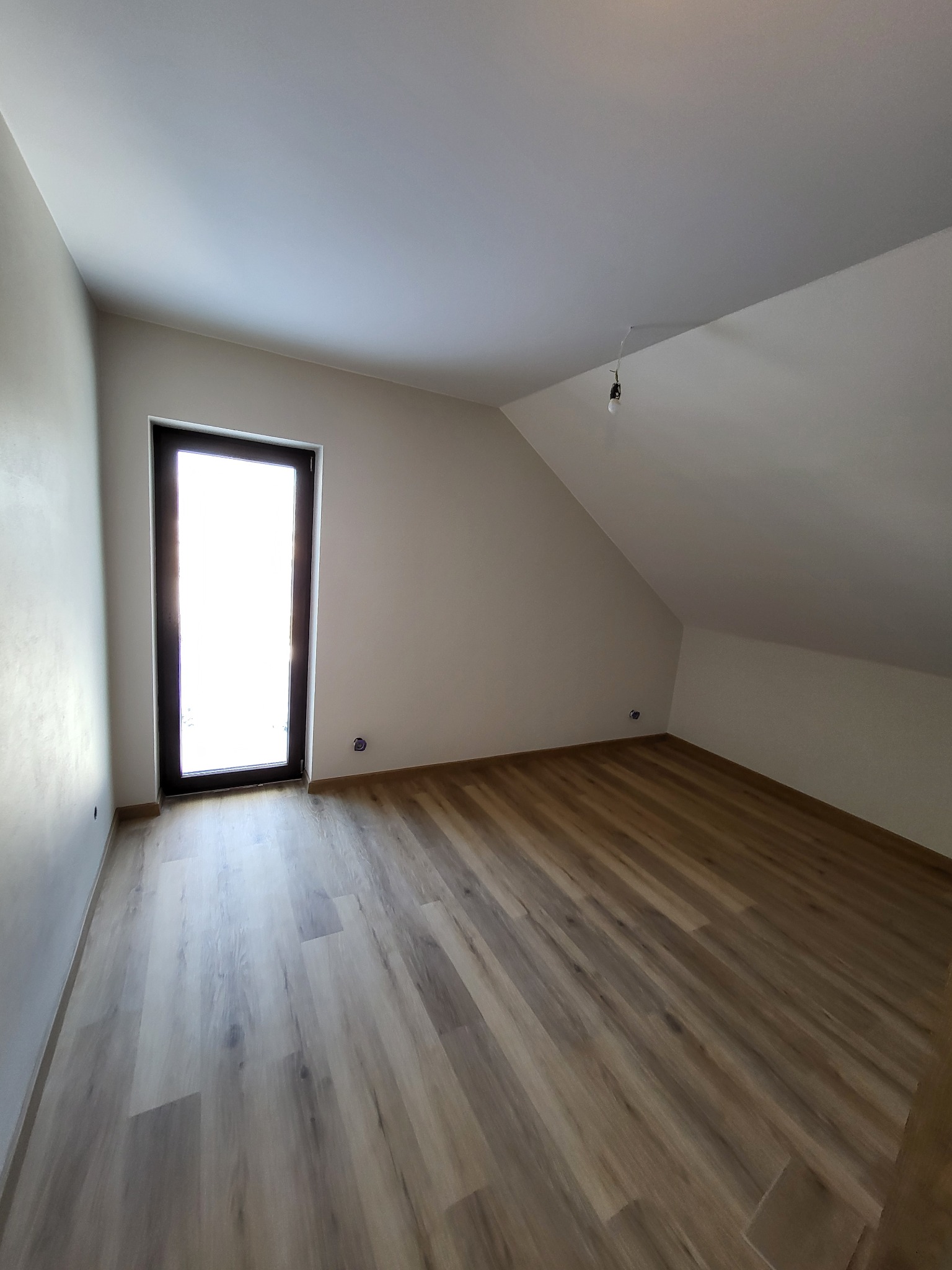 Poddasze po remoncie: świeżo ułożone panele podłogowe w odcieniach beżu i brązu, białe ściany z widocznymi gniazdkami elektrycznymi, okno z ciemną ramą wpuszczające jasne światło, surowa żarówka...