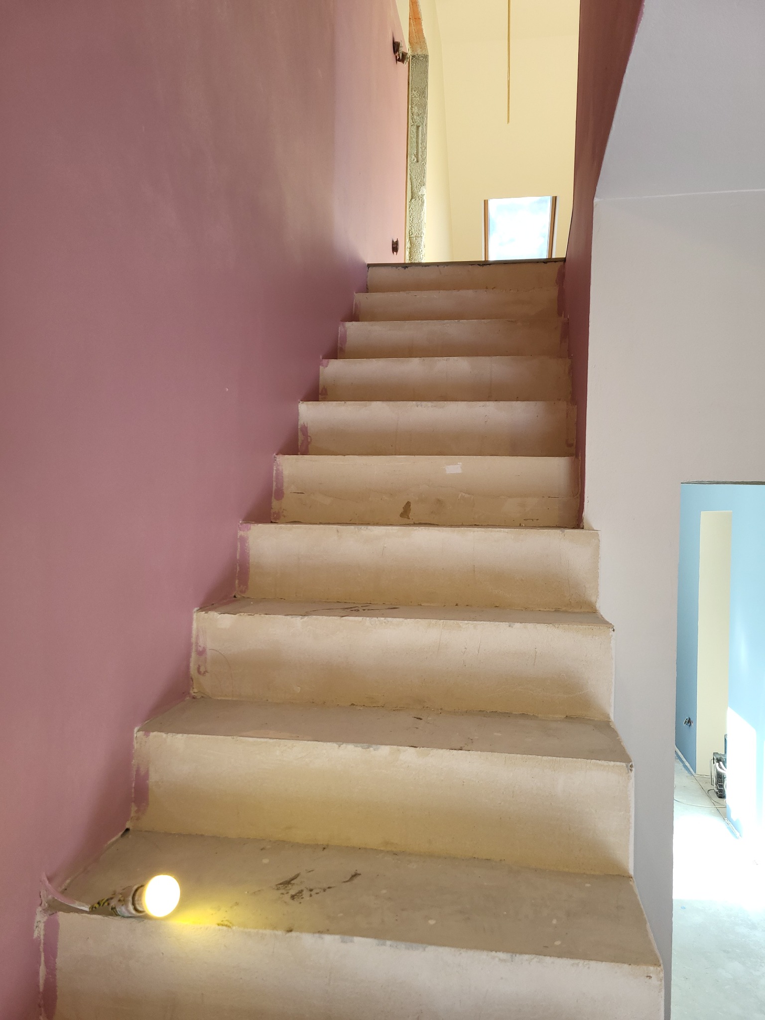 Schody w trakcie malowania, z widocznym tynkiem cw na ścianie i żarówką oświetlającą dolny stopień, w tle widoczne kolejne pomieszczenia w trakcie remontu.
