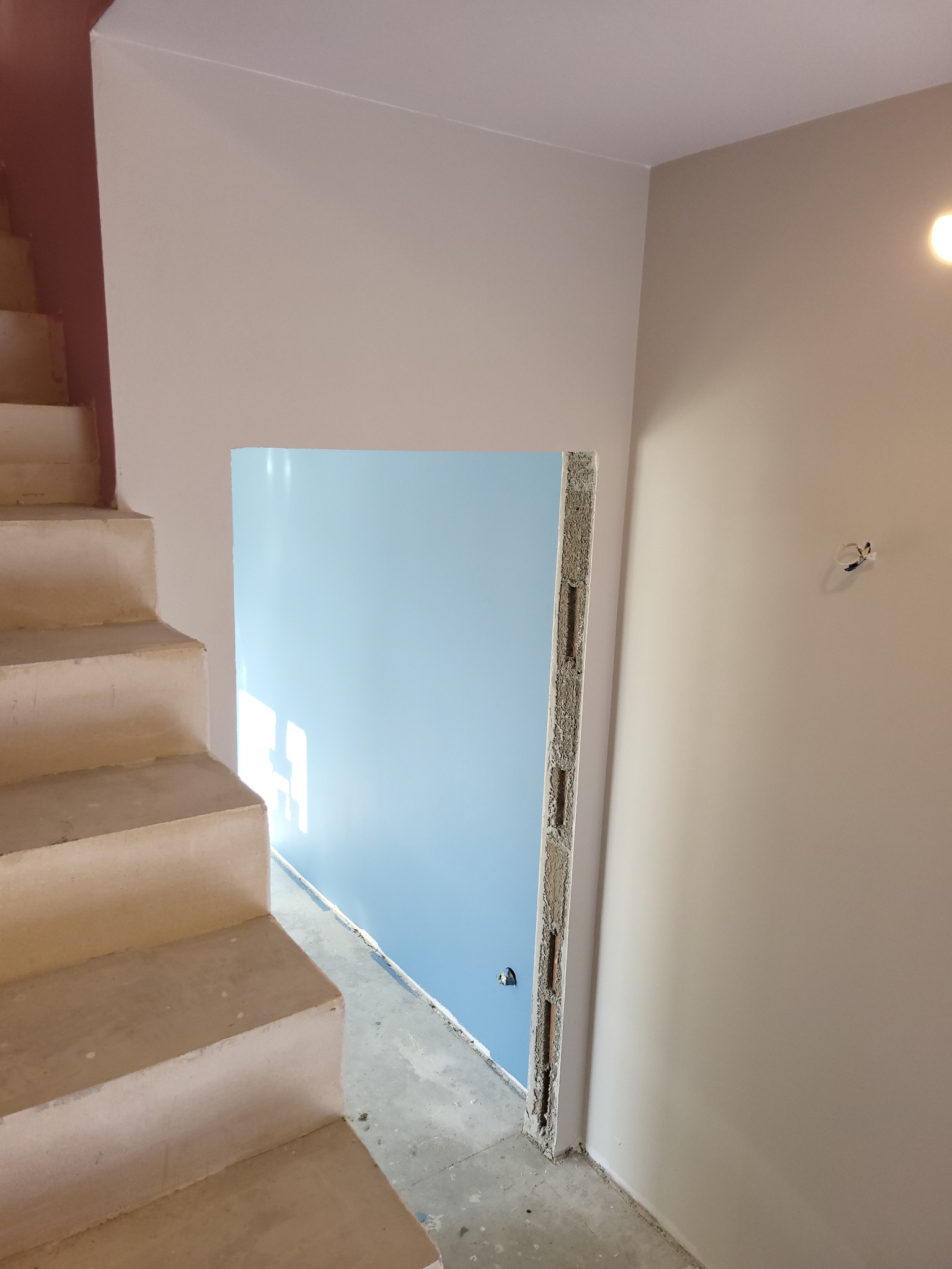 Wnętrze w trakcie prac wykończeniowych: widok na surowe schody, ścianę pomalowaną na beżowo oraz otwór drzwiowy z widoczną niebieską ścianą w tle.