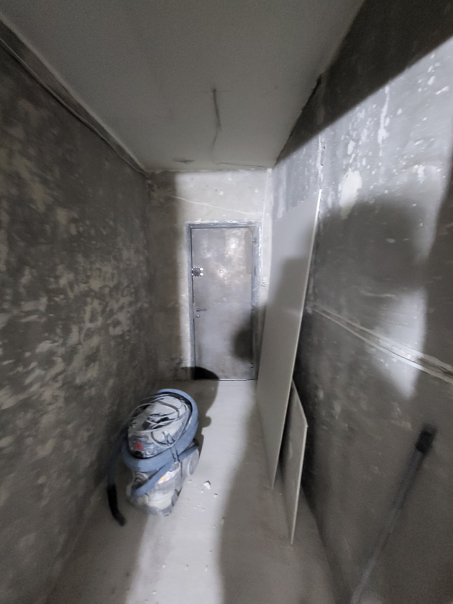 Wnętrze w trakcie prac: surowe ściany z widoczną strukturą, oparty o ścianę arkusz płyty karton-gips, odkurzacz przemysłowy w rogu, metalowe drzwi w tle.