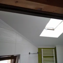 Biała łazienka z grzejnikiem drabinkowym na zielonej ścianie, widoczne białe płytki z teksturą, okno dachowe i drewniany element sufitu.