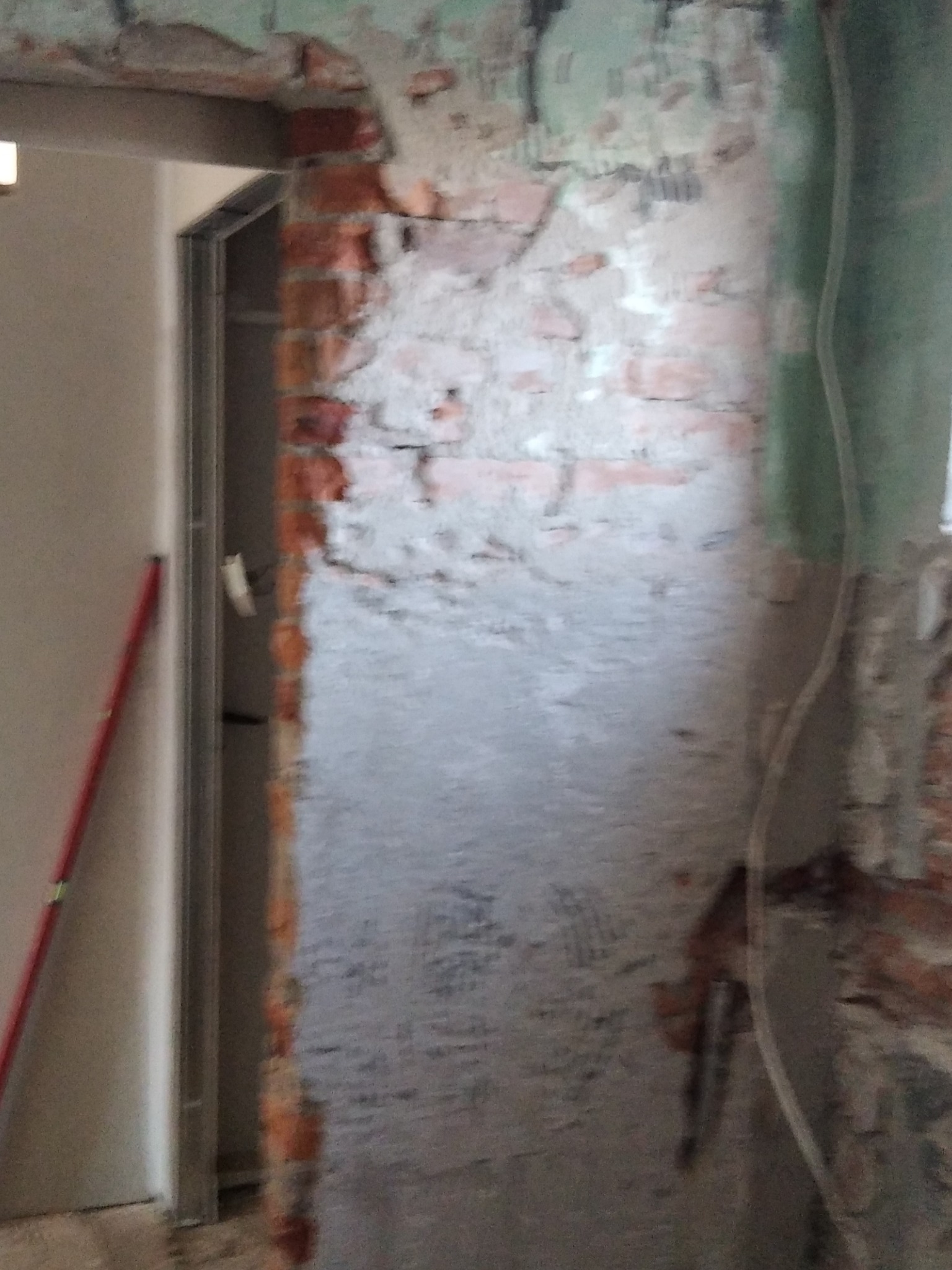 Niedokończone przejście w ścianie z widoczną czerwoną cegłą i resztkami starego tynku, przygotowywane do dalszych prac wykończeniowych, z widoczną łatą na ścianie i profilem metalowym ościeżnicy.