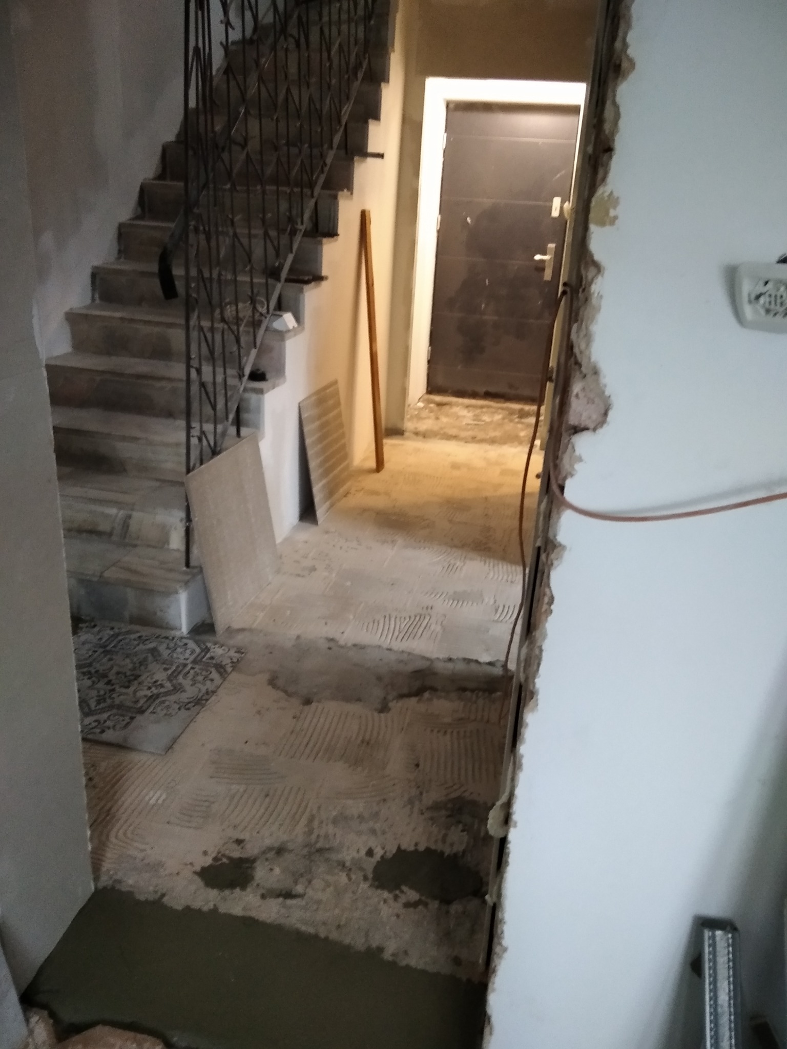 Klatka schodowa w trakcie remontu: widoczne przygotowane podłoże pod płytki, schody z metalową balustradą, fragment ściany z widocznym uszkodzeniem tynku, drzwi w tle.