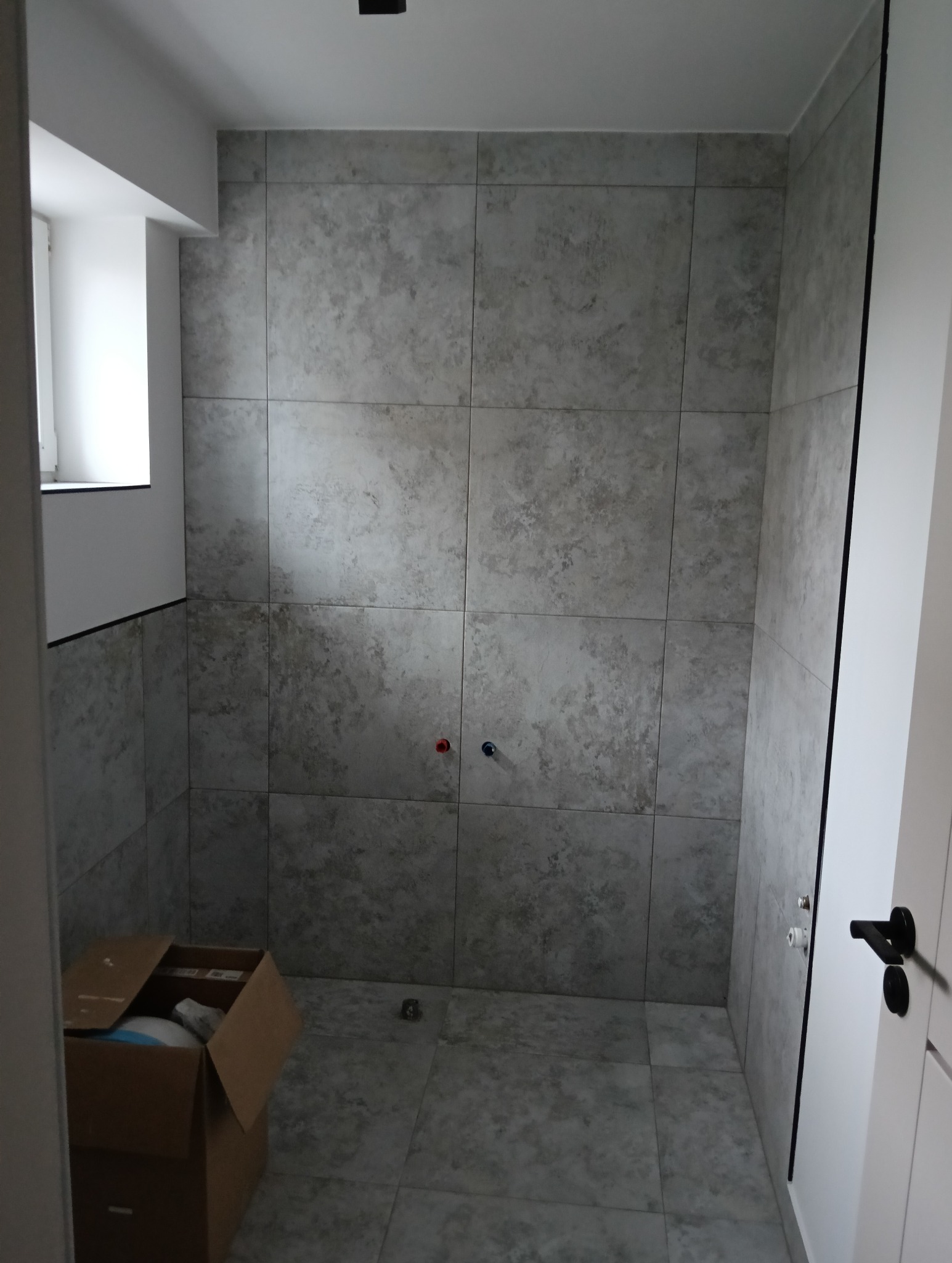 Wykończona łazienka z szarymi płytkami imitującymi kamień, widoczne przyłącza wodne, karton z akcesoriami w rogu pomieszczenia.