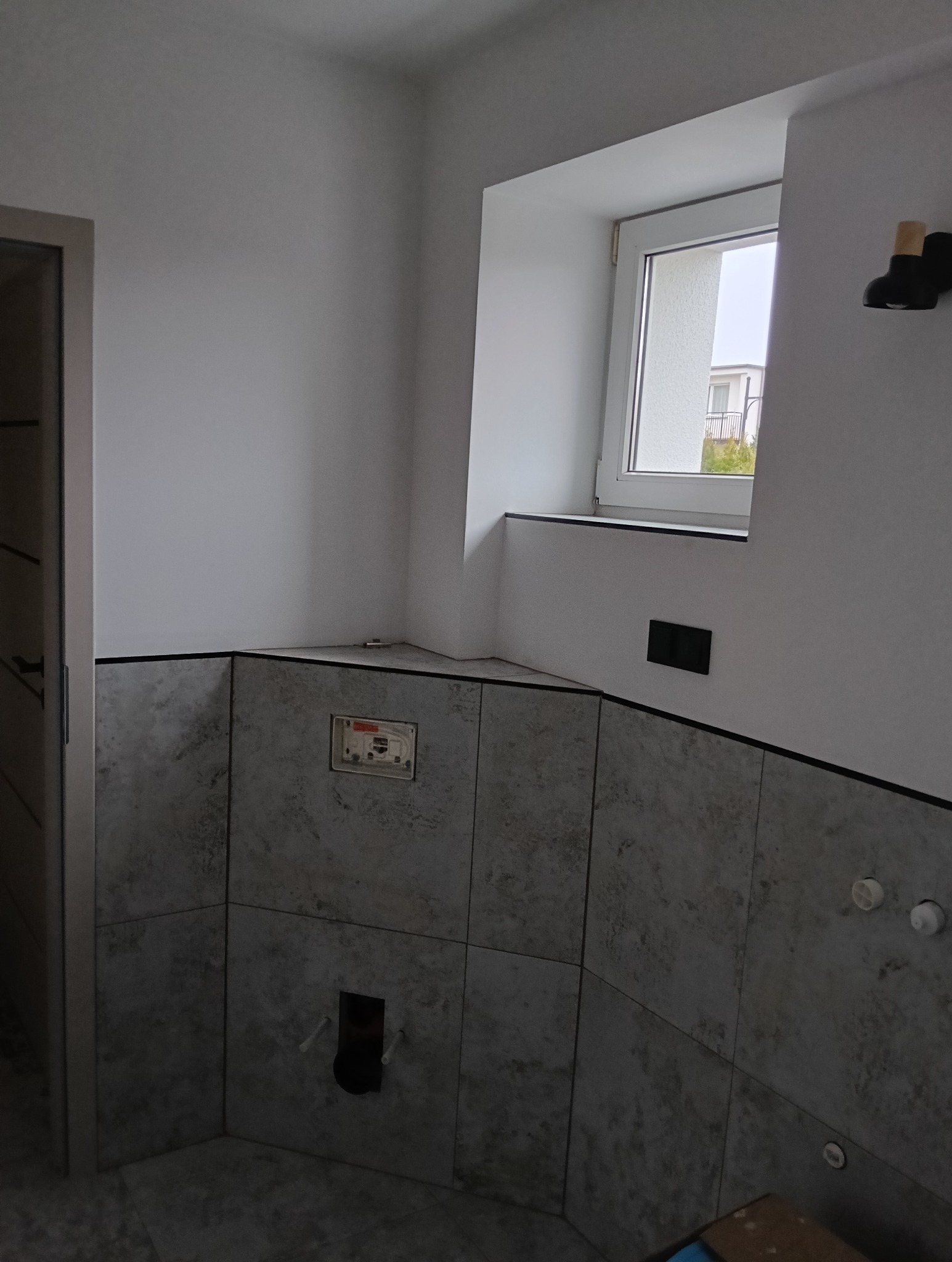 Wnętrze łazienki w Leśniewie w trakcie wykańczania, widoczne szare płytki na ścianach, okno z widokiem na zewnątrz oraz przygotowane instalacje pod armaturę.