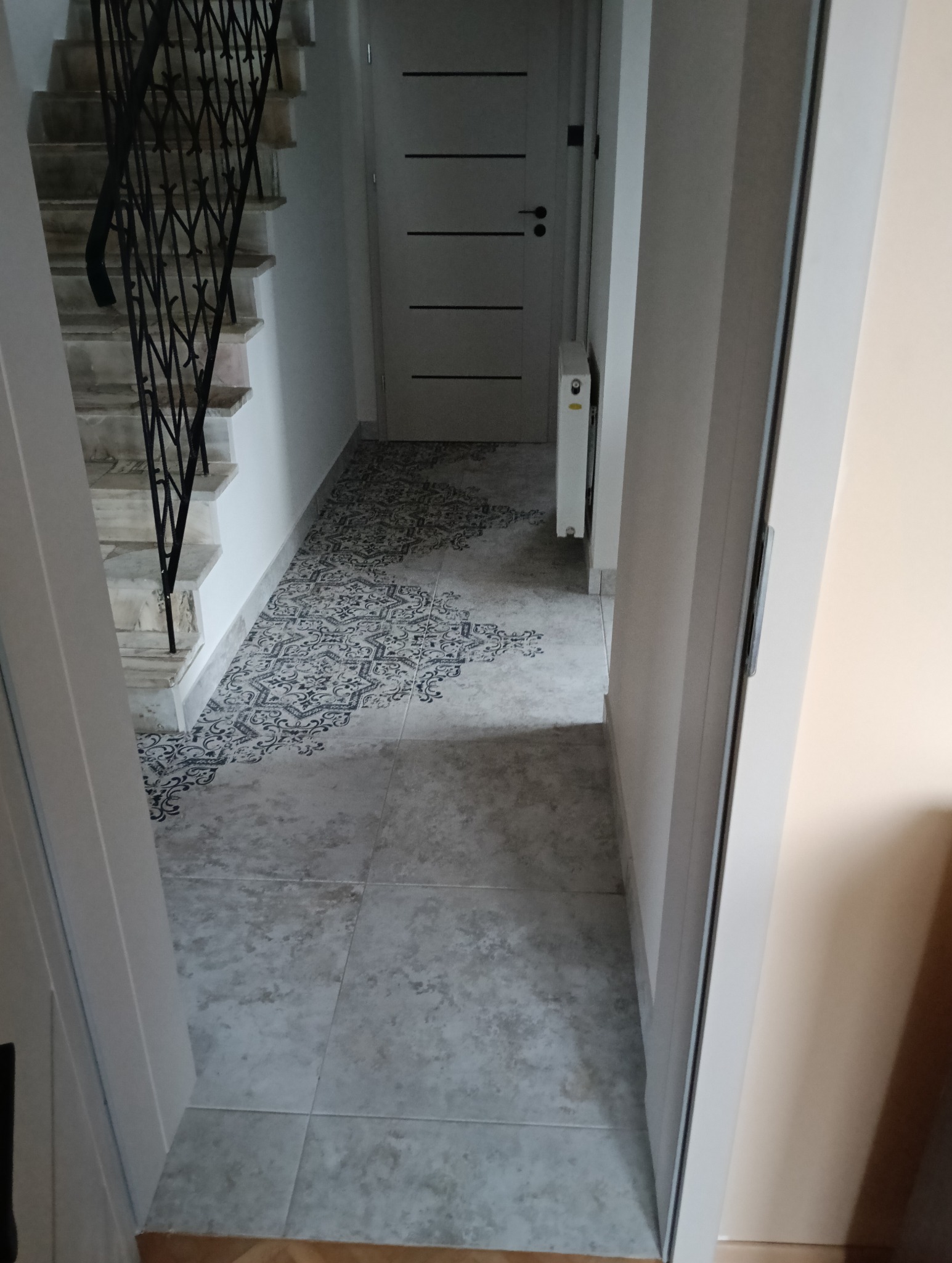 Korytarz z szarymi płytkami podłogowymi, dekoracyjnym wzorem w strefie wejściowej i schodami z czarną, metalową balustradą po lewej stronie.