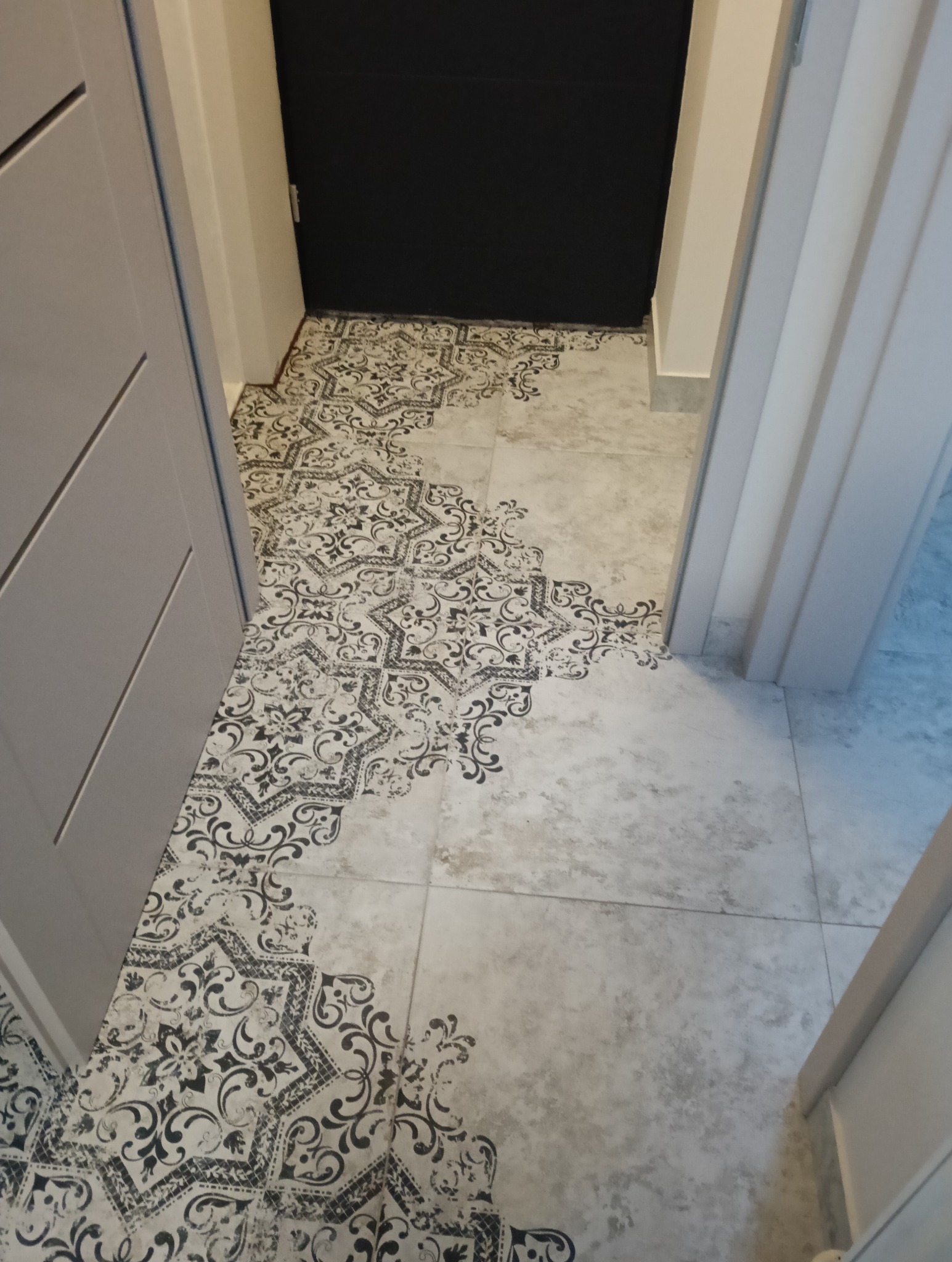 Wykończona podłoga w przejściu z wzorzystych, beżowo-czarnych płytek ceramicznych, widoczna czarna ściana i fragmenty białych ościeżnic drzwi.