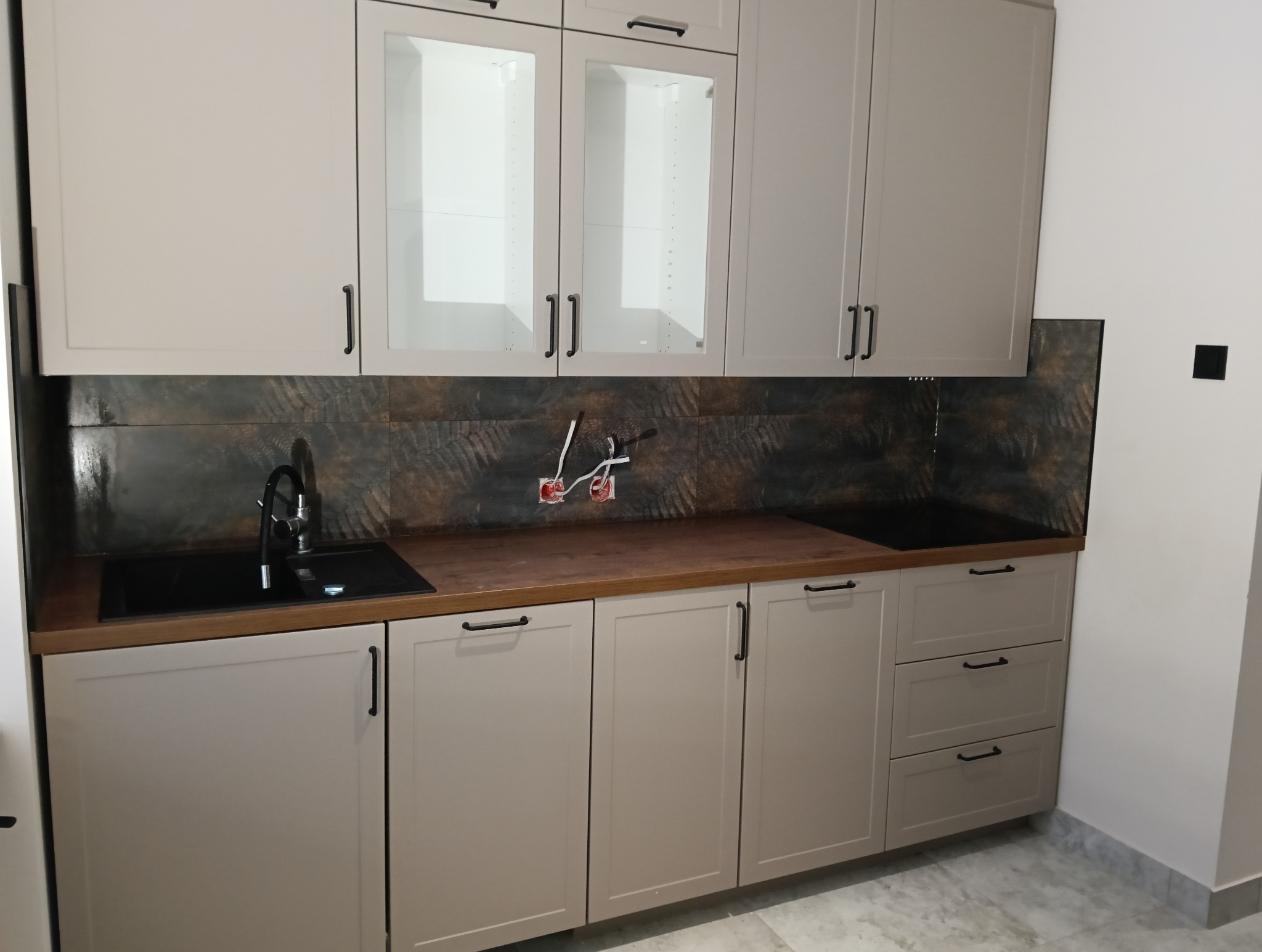 Kuchnia z beżowymi szafkami, czarnym zlewozmywakiem i brązową deską, widoczne gniazdka elektryczne przed montażem.