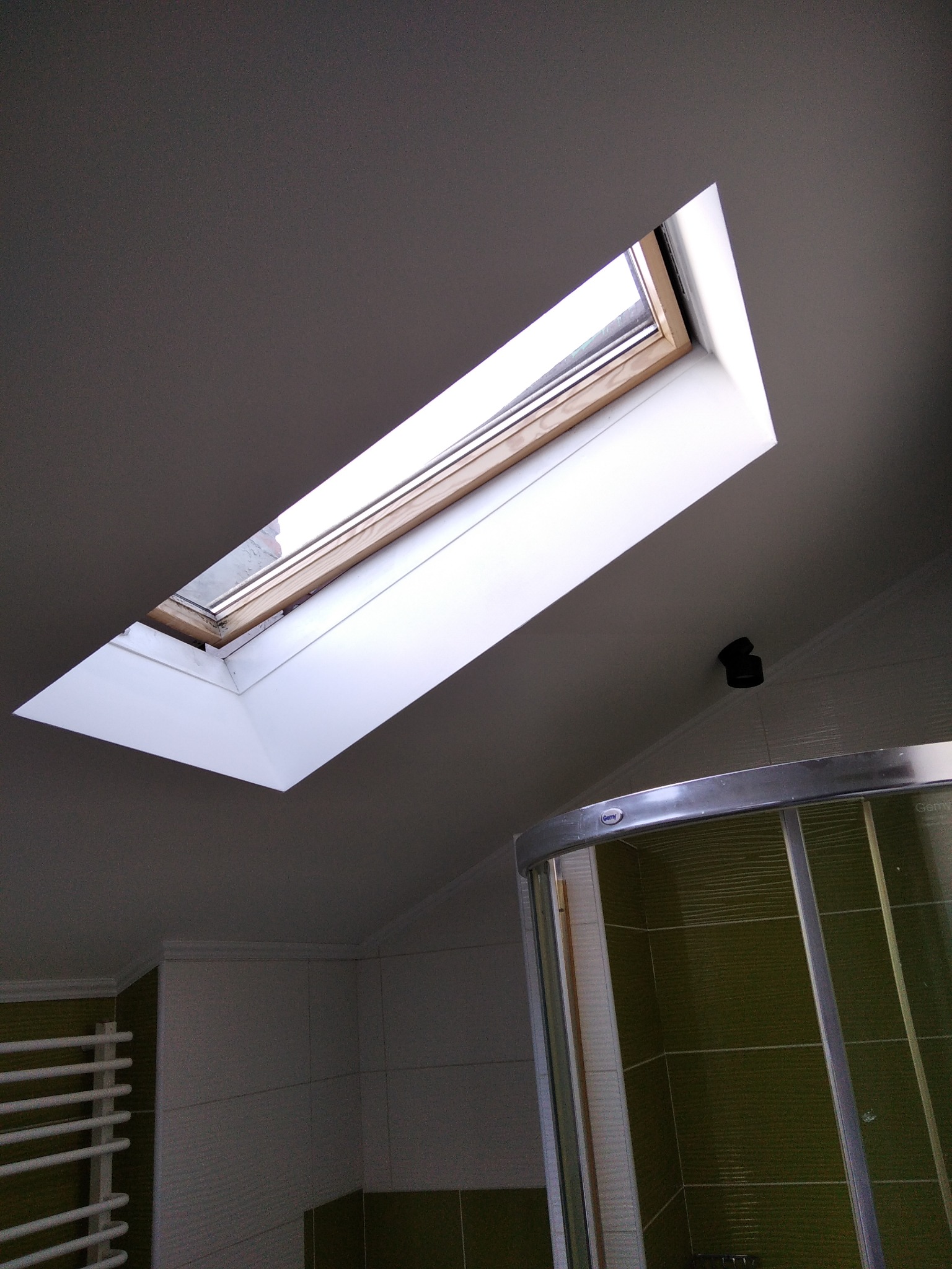 Wnętrze łazienki z oknem dachowym, widoczna kabina prysznicowa z zielonymi płytkami i grzejnik drabinkowy.