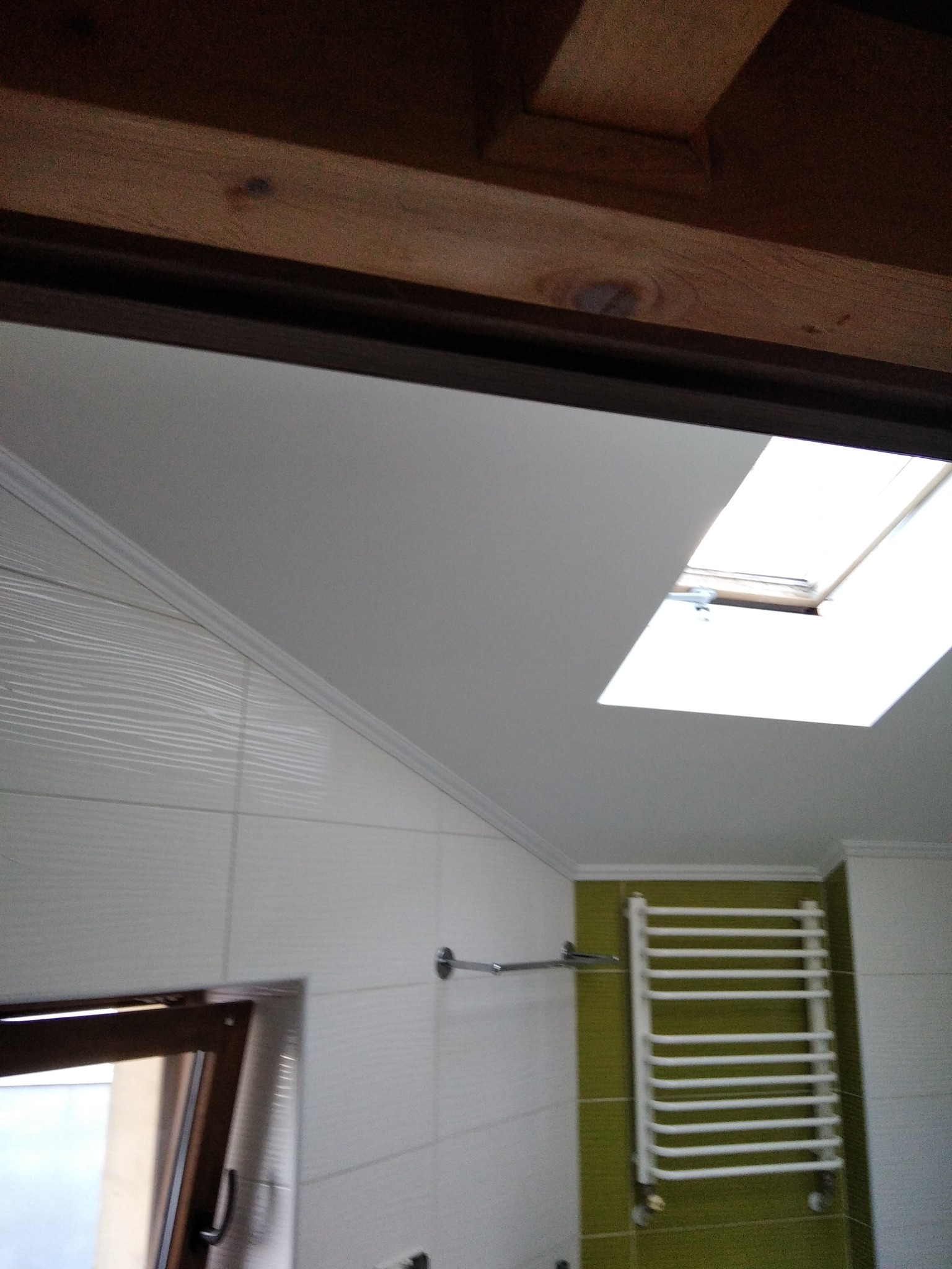 Biała łazienka z grzejnikiem drabinkowym na zielonej ścianie, widoczne białe płytki z teksturą, okno dachowe i drewniany element sufitu.