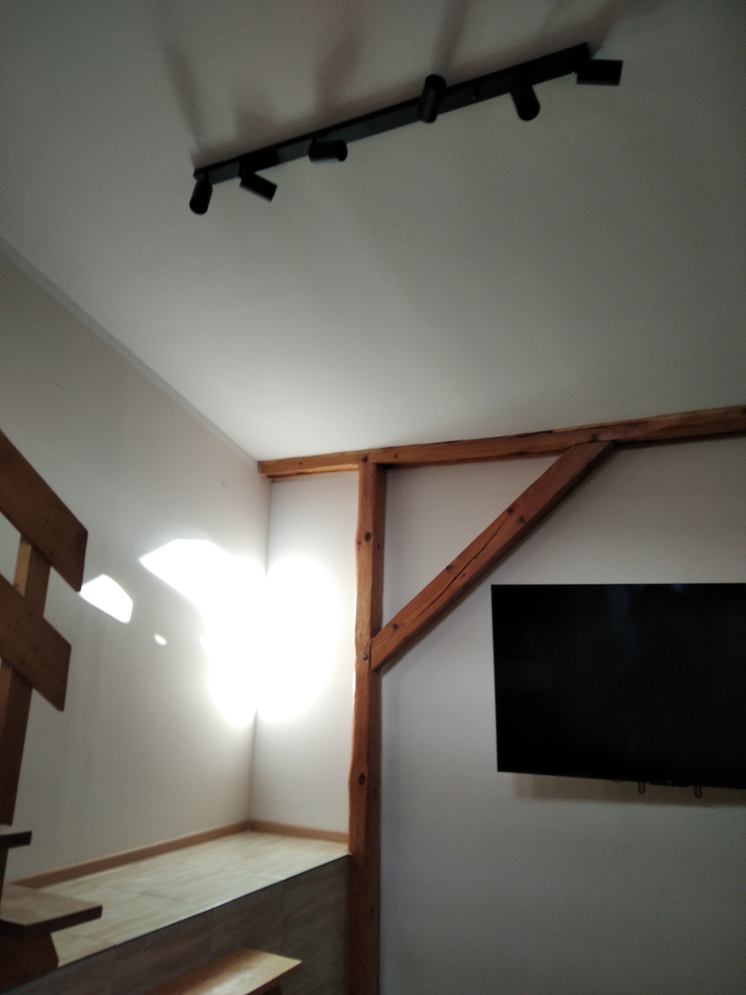 Wnętrze pokoju z drewnianą konstrukcją nośną, oświetleniem punktowym na suficie i zamontowanym telewizorem na ścianie.