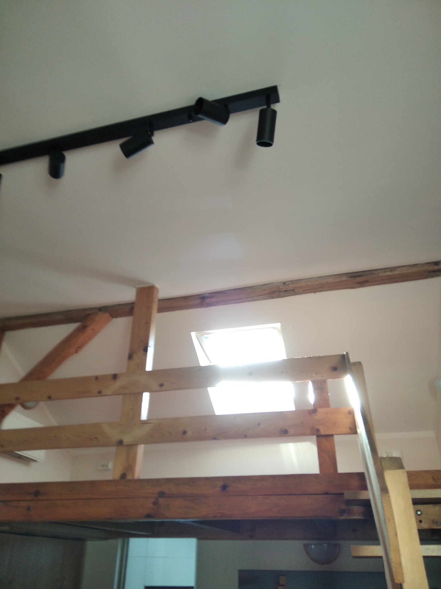 Drewniana konstrukcja antresoli ze schodami, widok z dołu, oświetlenie sufitowe na szynie i okno dachowe w tle.