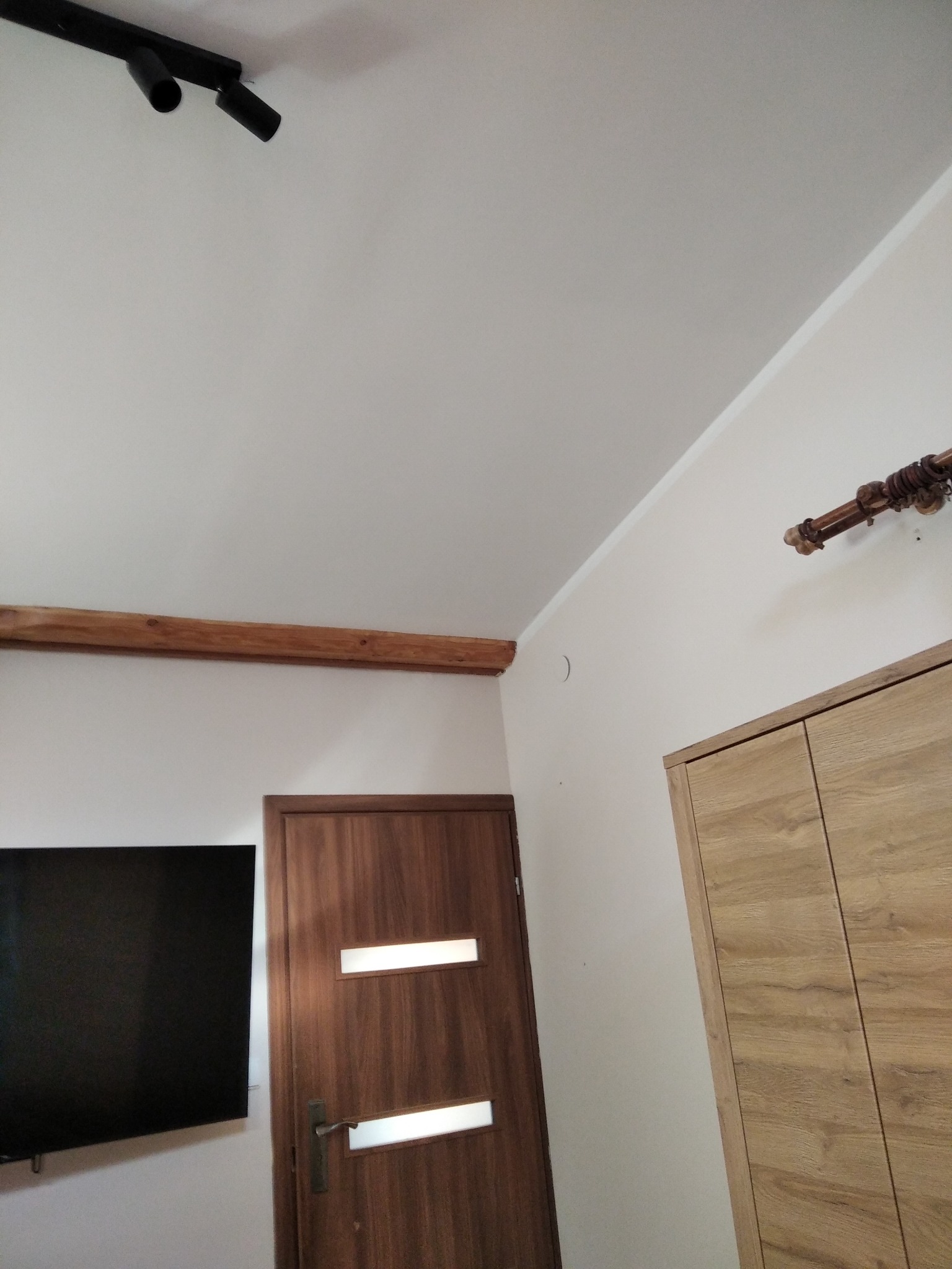 Wnętrze pokoju z drewnianą belką pod sufitem, brązowymi drzwiami z przeszkleniami, szafą z frontami imitującymi drewno i czarnym telewizorem na ścianie.