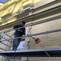 HOUSE-SERVICE Kamil Zieniuk - Pracownik na rusztowaniu czyści elewację budynku z cegły za pomocą specjalistycznej maszyny. Widoczne detale architektoniczne i okno. Jasna elewacja w trakcie czyszczenia.