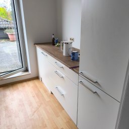 HOUSE-SERVICE Kamil Zieniuk - Białe szafki kuchenne z drewnianym blatem, puszka farby i akcesoria malarskie na blacie, widok na zewnątrz przez okno. Przygotowania do malowania.