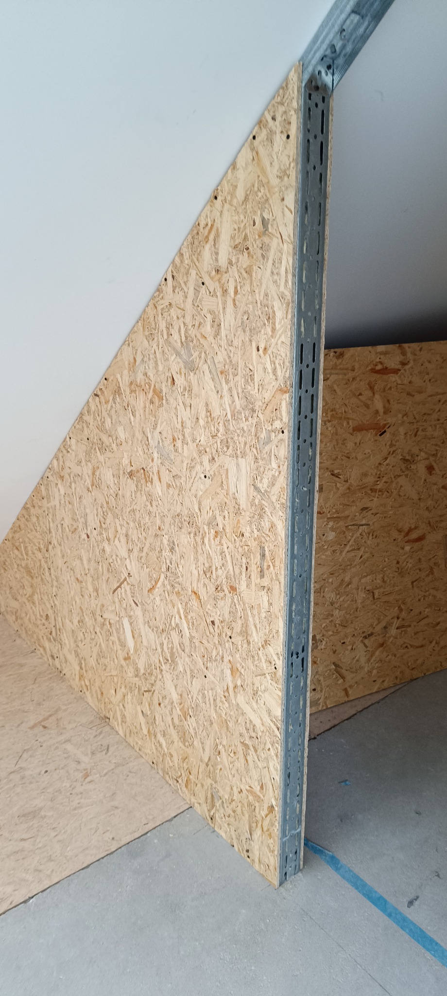 Szkieletowa konstrukcja ścianki działowej z płyty OSB, wzmocniona metalowym profilem, w surowym wnętrzu poddasza. Widoczne mocowania i fragment podłogi.