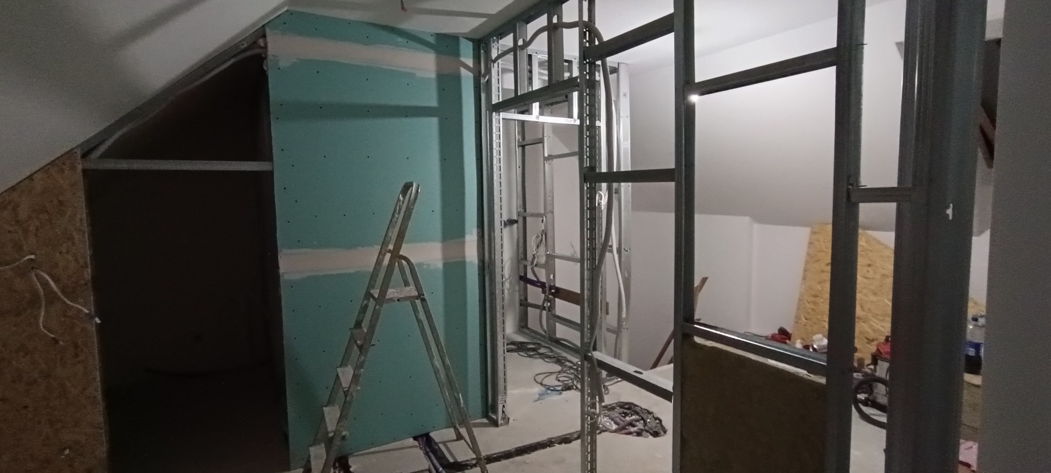 Szkielet ścianki działowej z profili stalowych i płyty gipsowej na poddaszu, widoczna drabina, przewody. Etap prac wykończeniowych.