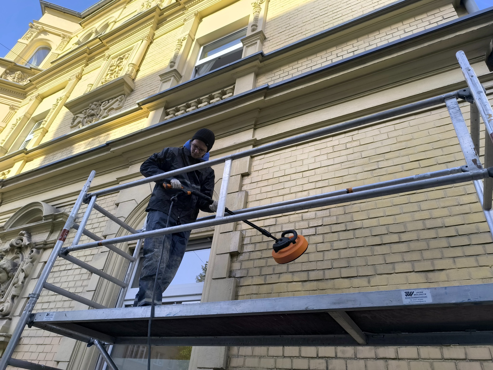 Pracownik na rusztowaniu czyści elewację budynku z cegły za pomocą specjalistycznej maszyny. Widoczne detale architektoniczne i okno. Jasna elewacja w trakcie czyszczenia.