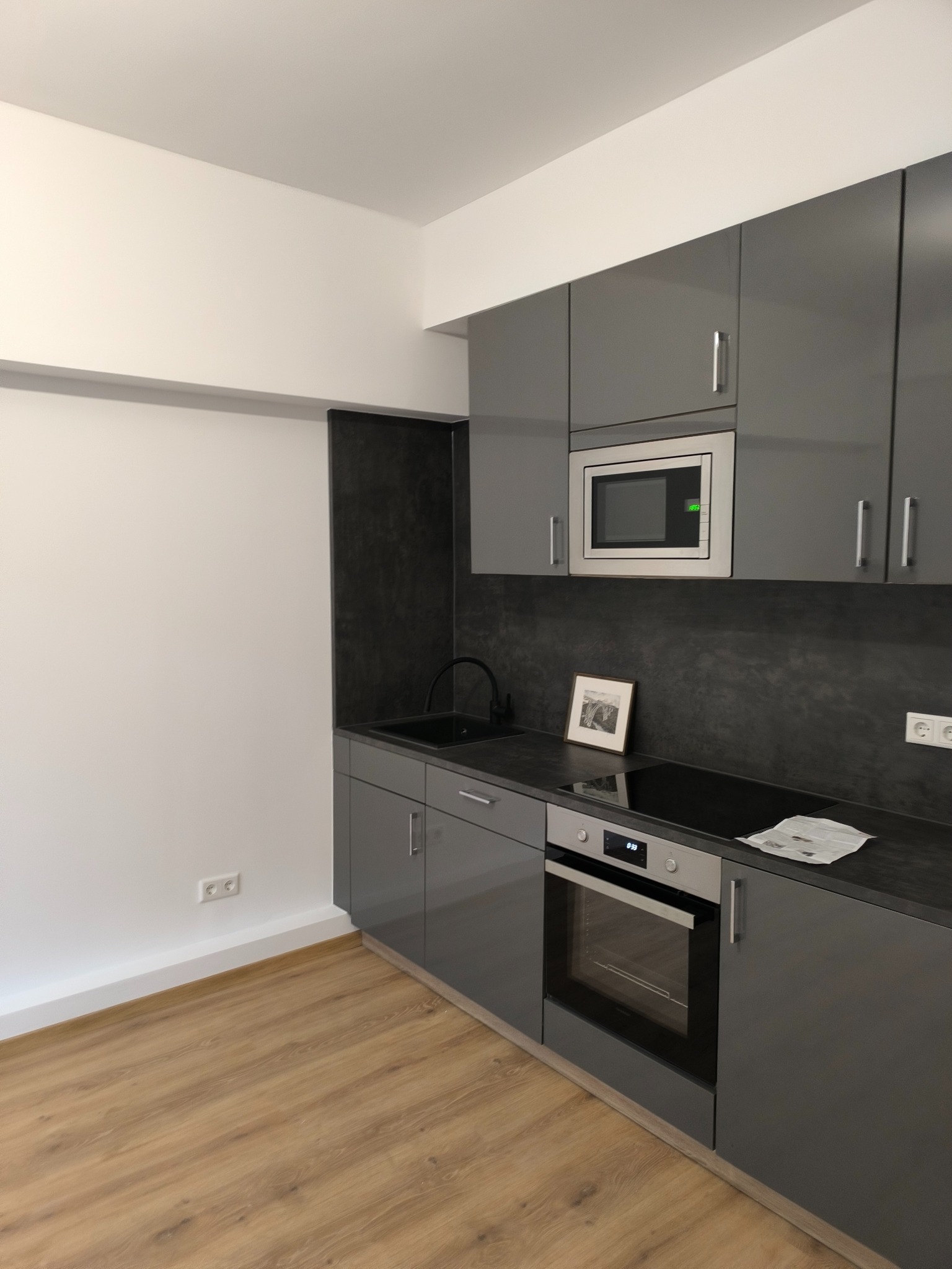 Nowoczesna, szara kuchnia z połyskiem, z wbudowanym piekarnikiem i mikrofalówką. Na blacie czarna płyta indukcyjna i ramka ze zdjęciem. Jasna podłoga z paneli.