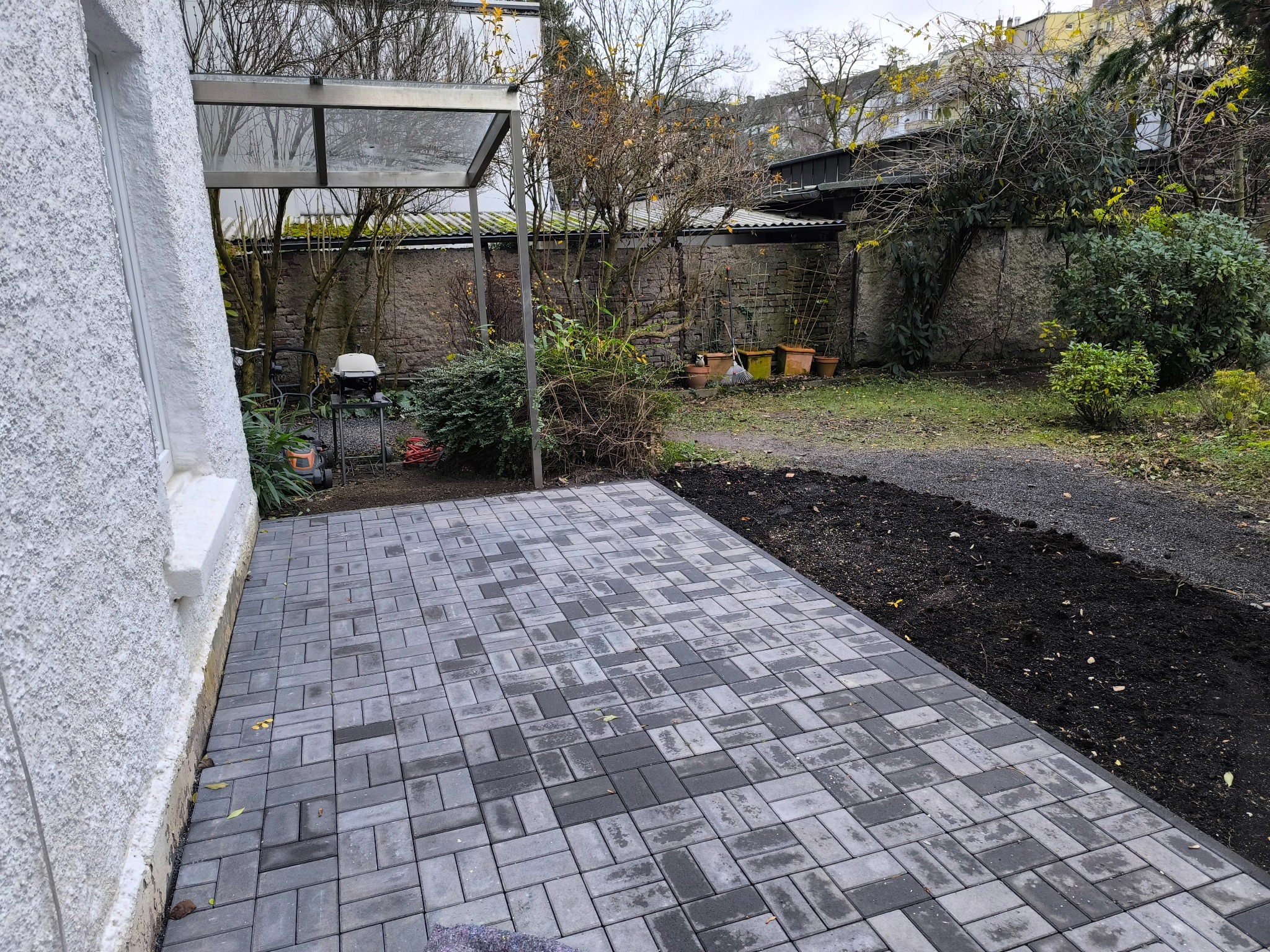 Ułożona szara kostka brukowa na tarasie przydomowym, z widocznym fragmentem ogrodu i elementami architektury ogrodowej w tle. Widok z perspektywy narożnej.