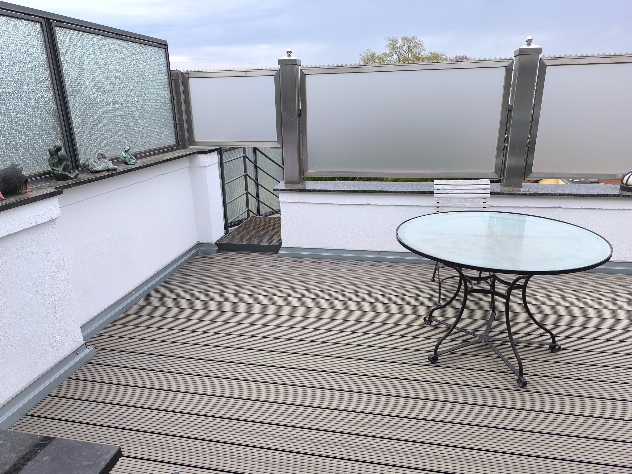 Nowoczesny taras na dachu z drewnianą podłogą, szklanymi balustradami i metalowymi słupkami. Na tarasie okrągły stół z blatem ze szkła i metalowe krzesło.