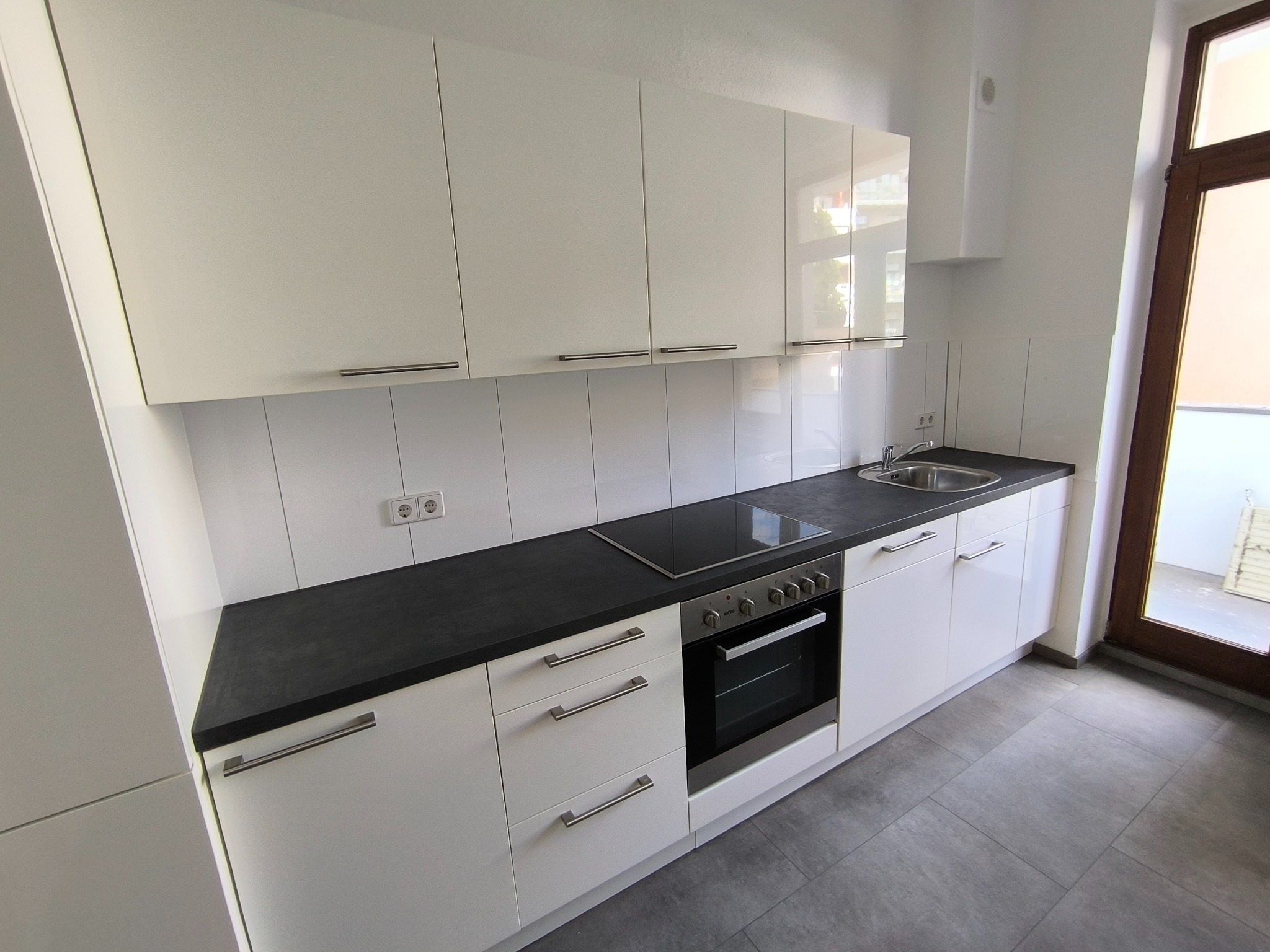 Nowoczesna, biała kuchnia z czarnym blatem i płytą indukcyjną. Minimalistyczny design, szare płytki podłogowe, okno z widokiem na balkon. Ukończona realizacja.