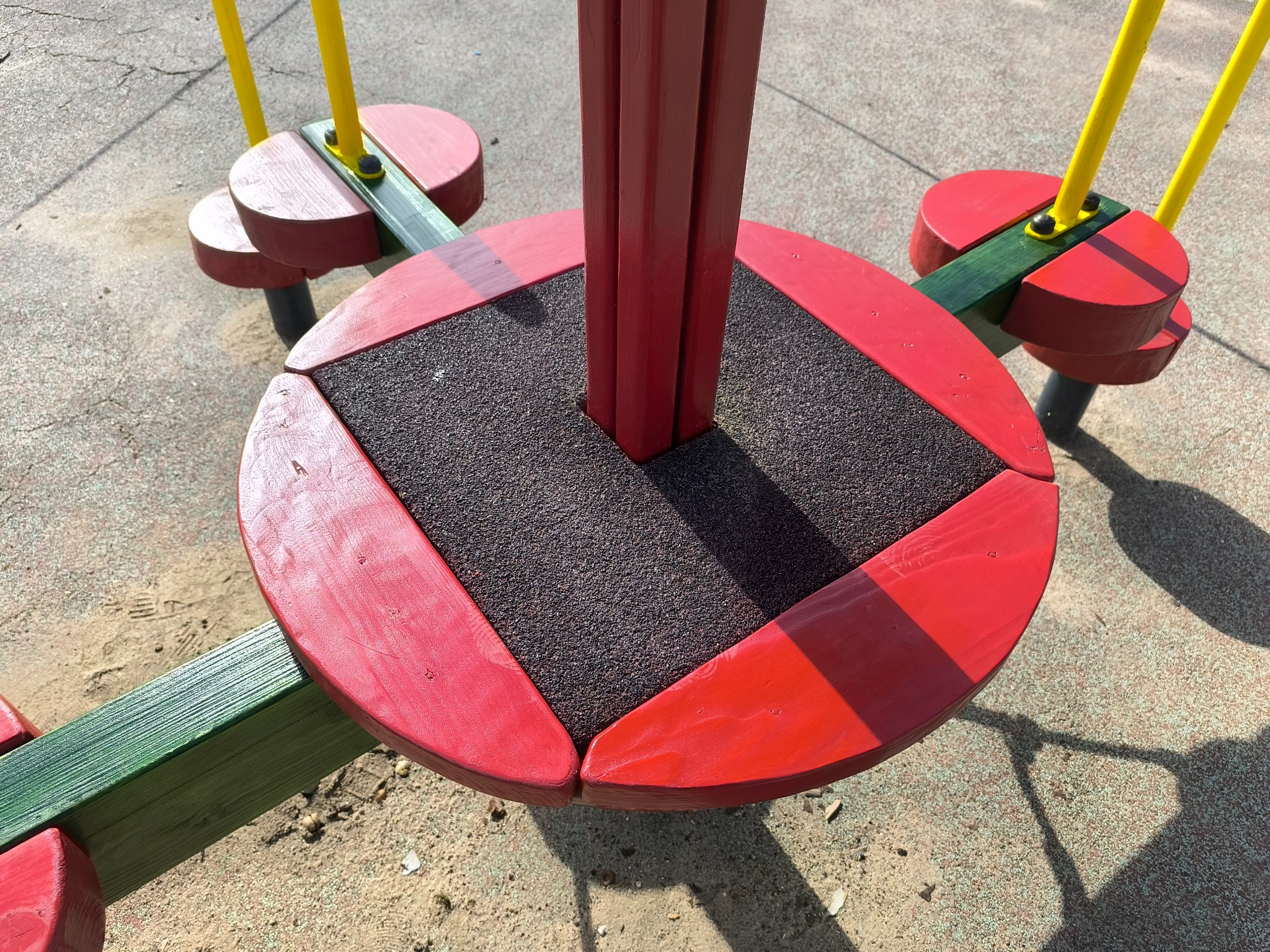 Kolorowy karuzela na placu zabaw z czerwonymi i zielonymi elementami drewnianymi oraz czarną gumową powierzchnią do siedzenia, oświetlona naturalnym światłem słonecznym.