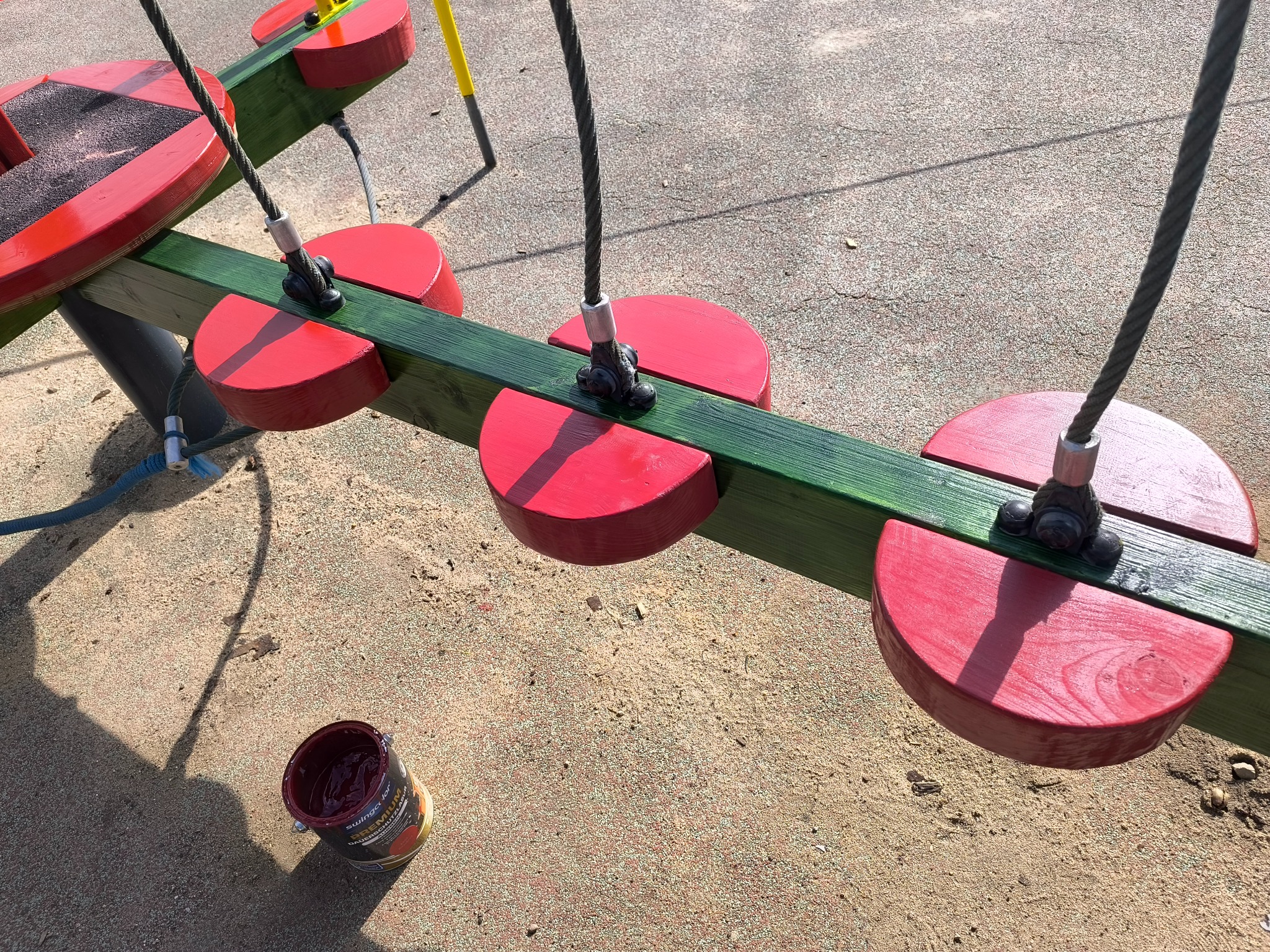 Malowanie drewnianego placu zabaw na zewnątrz. Widoczna puszka z farbą, pomalowane elementy na czerwono i zielono. Detal z bliska, etap prac.