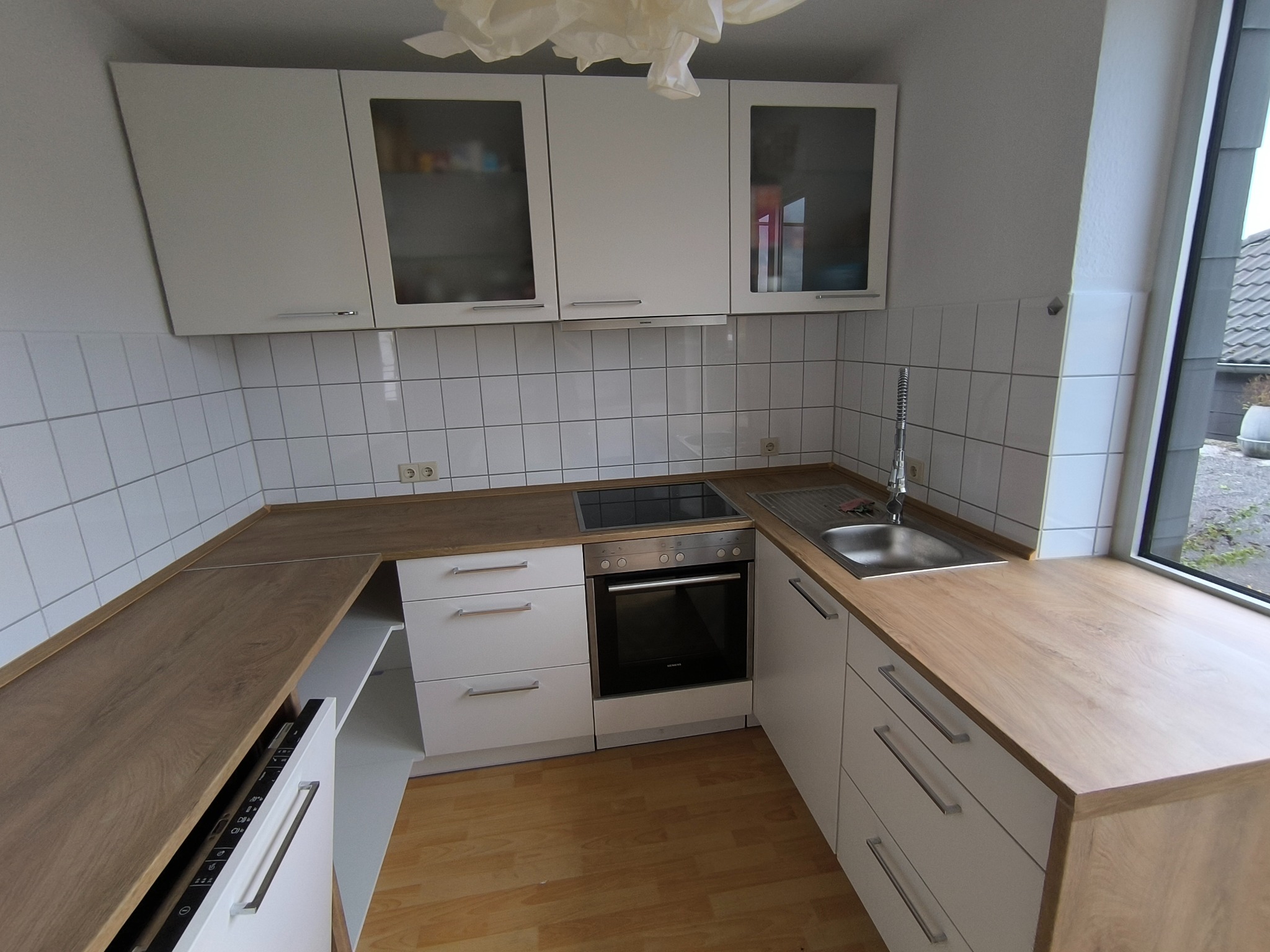 Nowoczesna, jasna kuchnia z białymi szafkami i drewnianym blatem. Widoczny zlew, płyta grzewcza i okno z widokiem na zewnątrz. Minimalistyczny design.