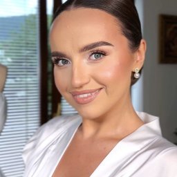 Astaaroth Beauty - Portret kobiety z subtelnym makijażem, upiętymi włosami i perłowymi kolczykami na tle okna z widokiem na góry. Delikatny uśmiech.