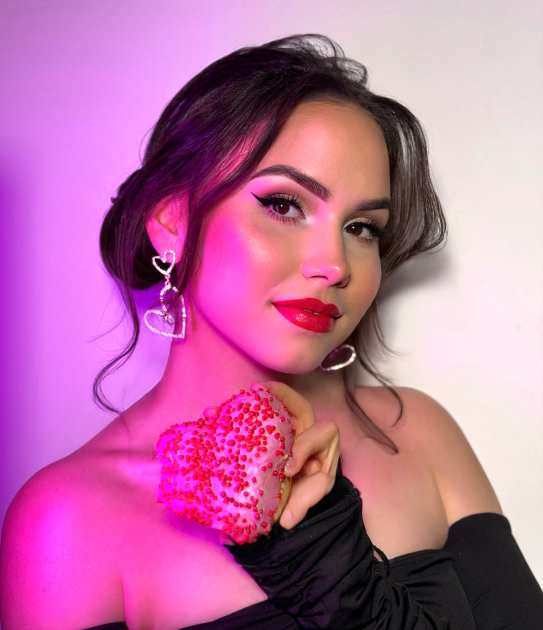 Portret kobiety z wyrazistym makijażem w Tymbarku, trzymającej pączka w kształcie serca z czerwonymi posypkami, w scenerii z fioletowym oświetleniem i kolczykami w kształcie serc.