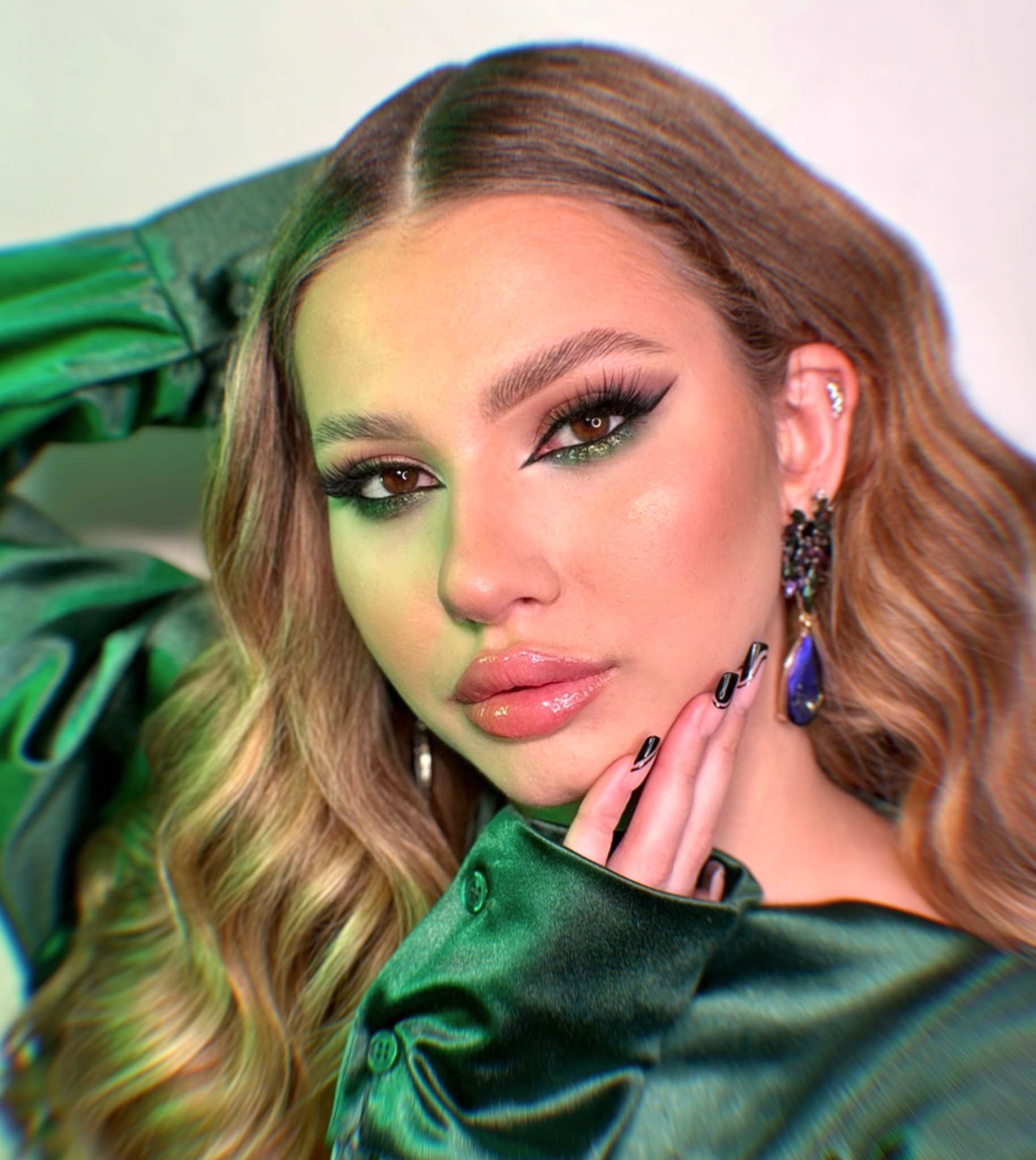 Portret kobiety z profesjonalnym makijażem oczu w odcieniach zieleni, błyszczącymi ustami i manicure z czarnymi paznokciami, pozującej w zielonej satynowej koszuli i z kolczykiem w chrząstce ucha.