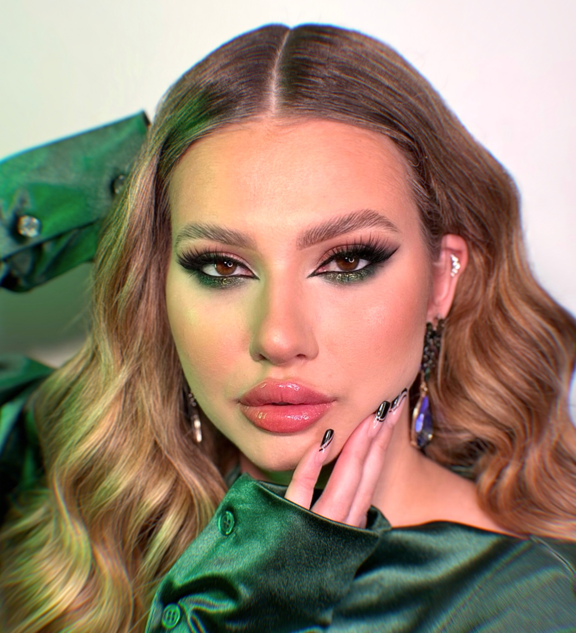 Portret kobiety z profesjonalnym makijażem oczu w odcieniach zieleni i błyszczącymi ustami, stylizacją włosów w fale i manicure z czarnymi zdobieniami, ubrana w satynową, zieloną koszulę.