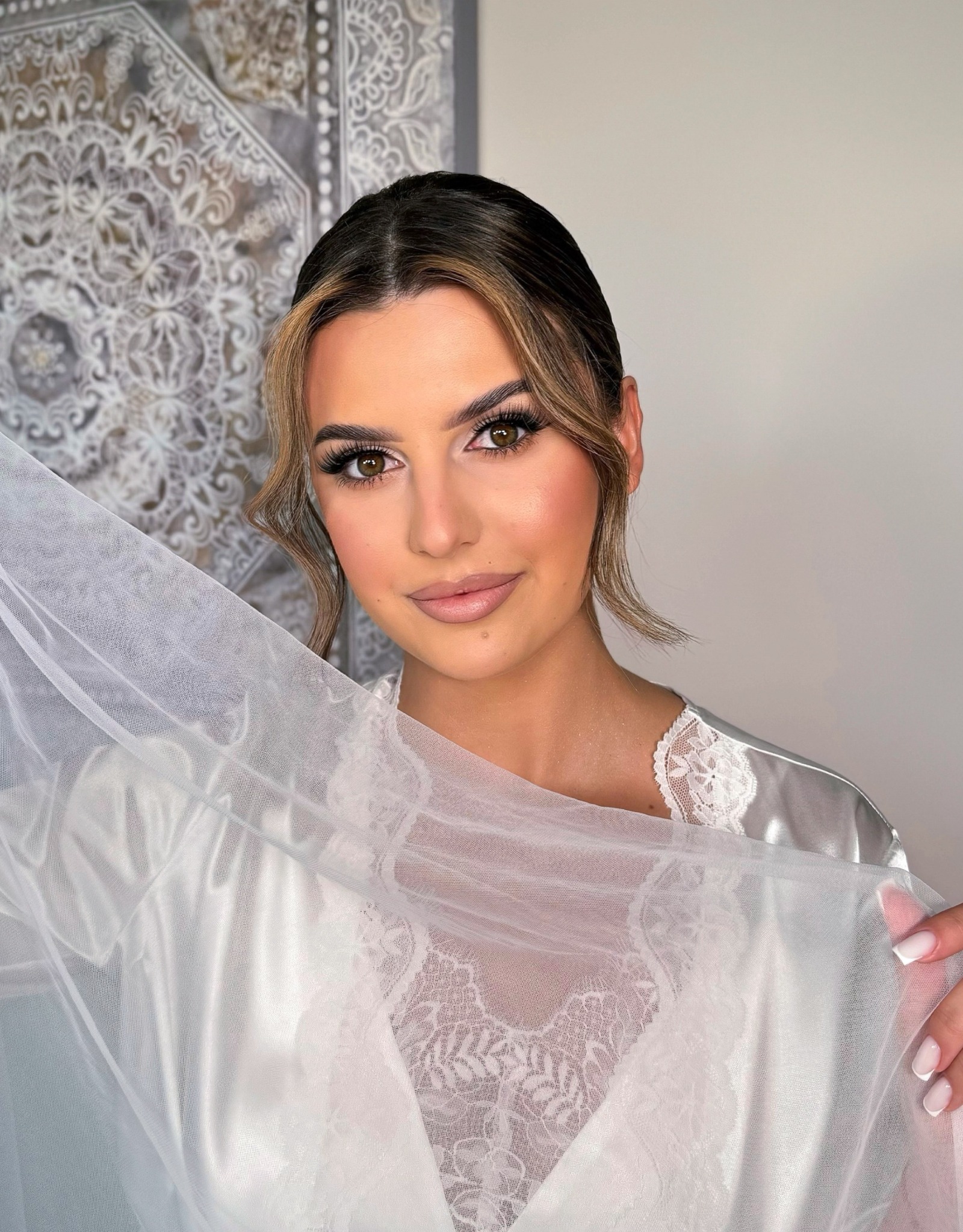 Portret kobiety z profesjonalnym makijażem i upiętymi włosami, trzymającej delikatną, białą tkaninę na tle wzorzystej ściany.