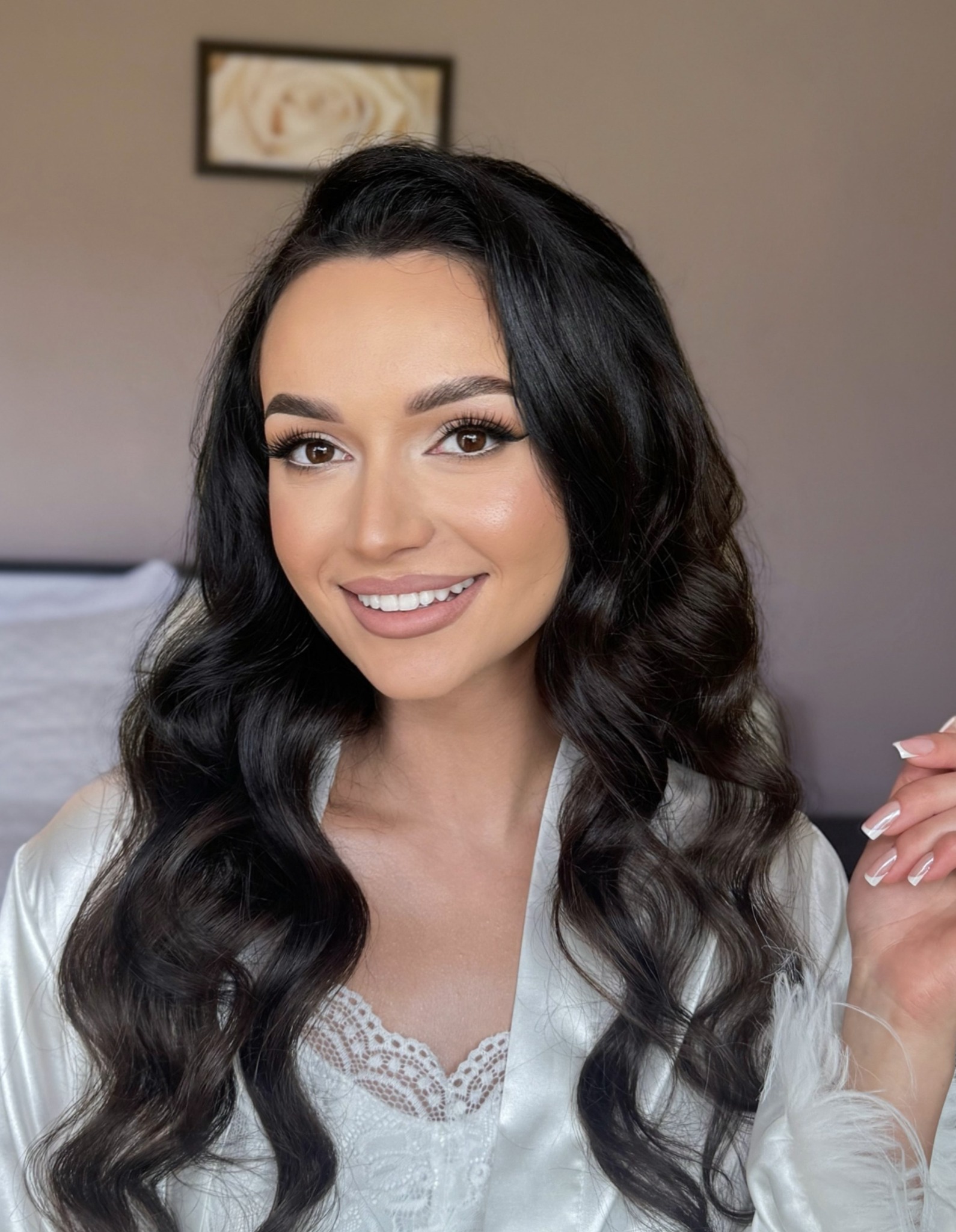 Uśmiechnięta brunetka w satynowym szlafroku prezentuje makijaż i fryzurę z ciemnymi, falowanymi włosami. Widoczny manicure francuski.