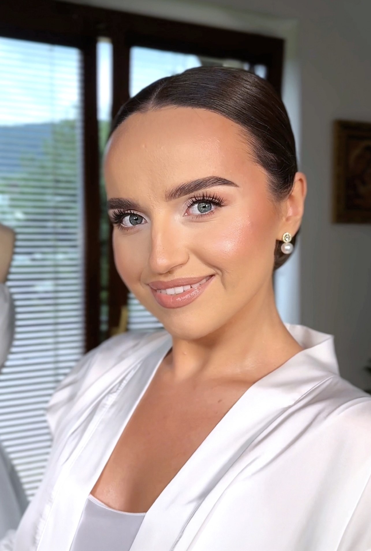 Portret kobiety z subtelnym makijażem, upiętymi włosami i perłowymi kolczykami na tle okna z widokiem na góry. Delikatny uśmiech.