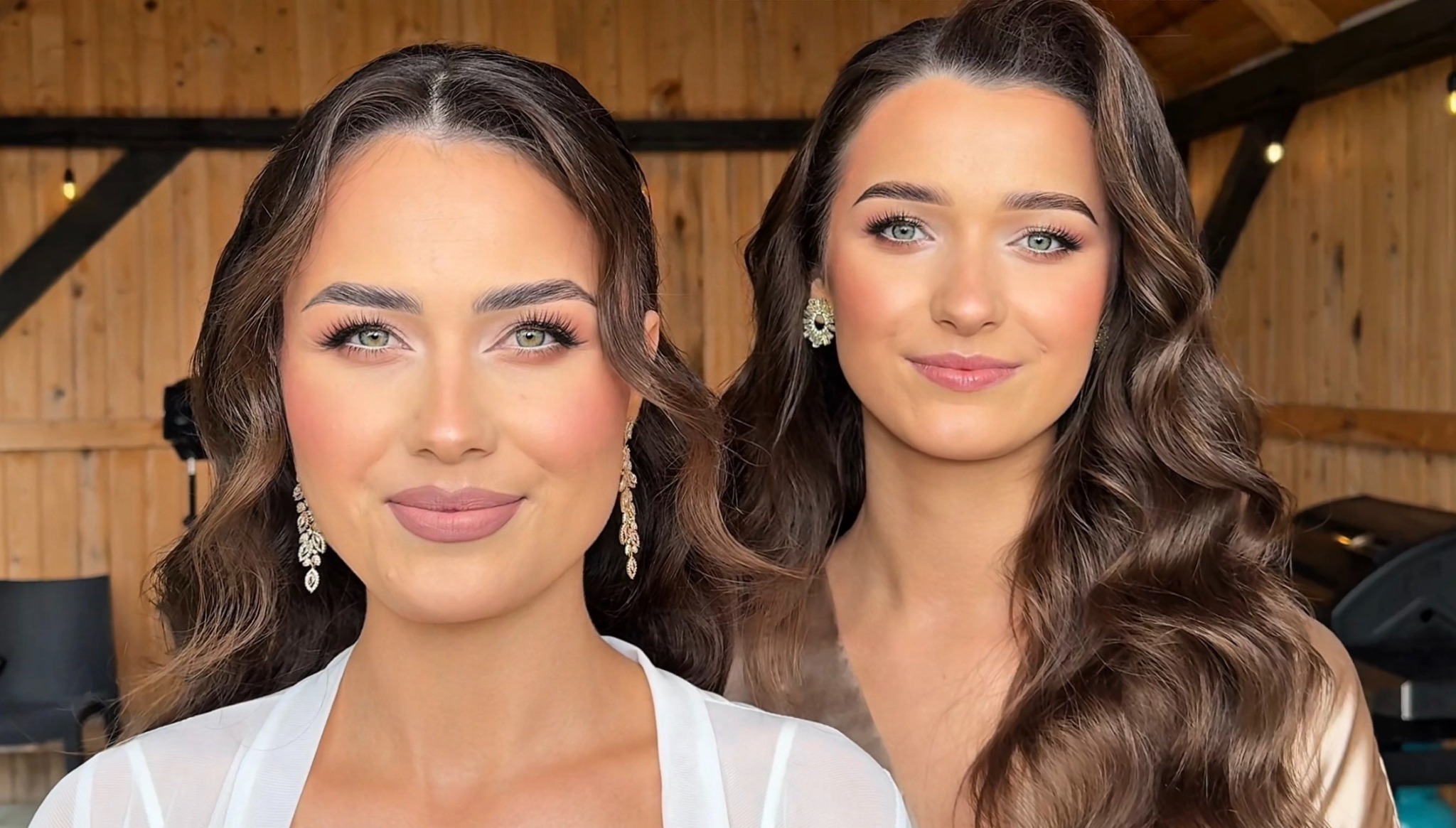 Portret dwóch uśmiechniętych kobiet z profesjonalnym makijażem i upiętymi, brązowymi włosami, pozujących na tle drewnianej ściany z oświetleniem.