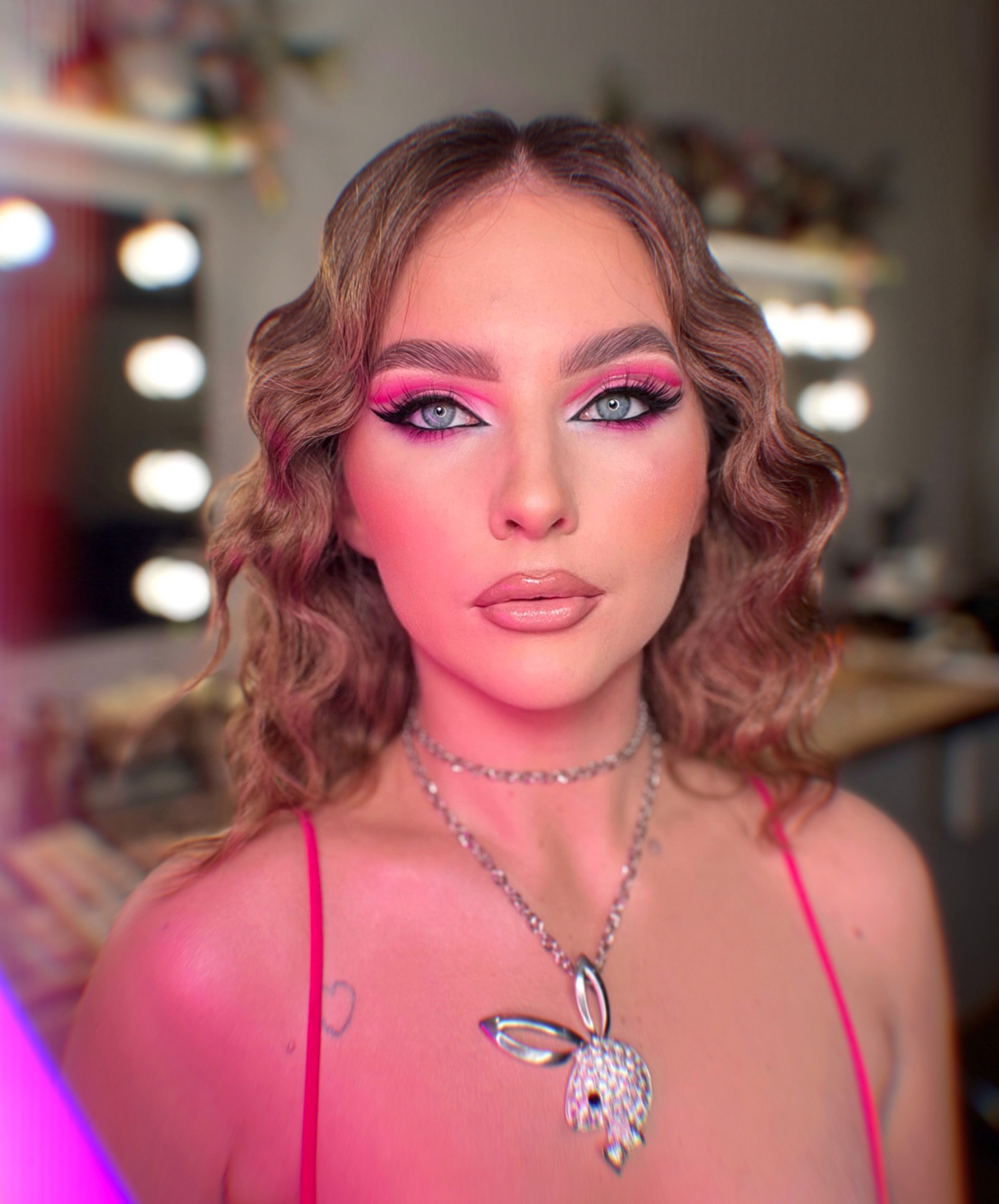 Portret kobiety z mocnym, różowym makijażem oczu, błyszczącymi ustami i delikatnymi falami na włosach, pozującej na tle lustra z oświetleniem