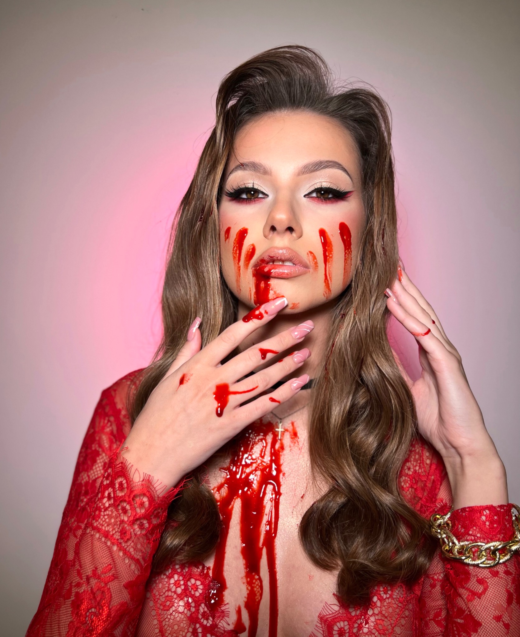 Portret kobiety z artystycznym makijażem imitującym krew, czerwona koronka na ubraniu, złota bransoleta, profesjonalny makijaż oczu z eyelinerem.