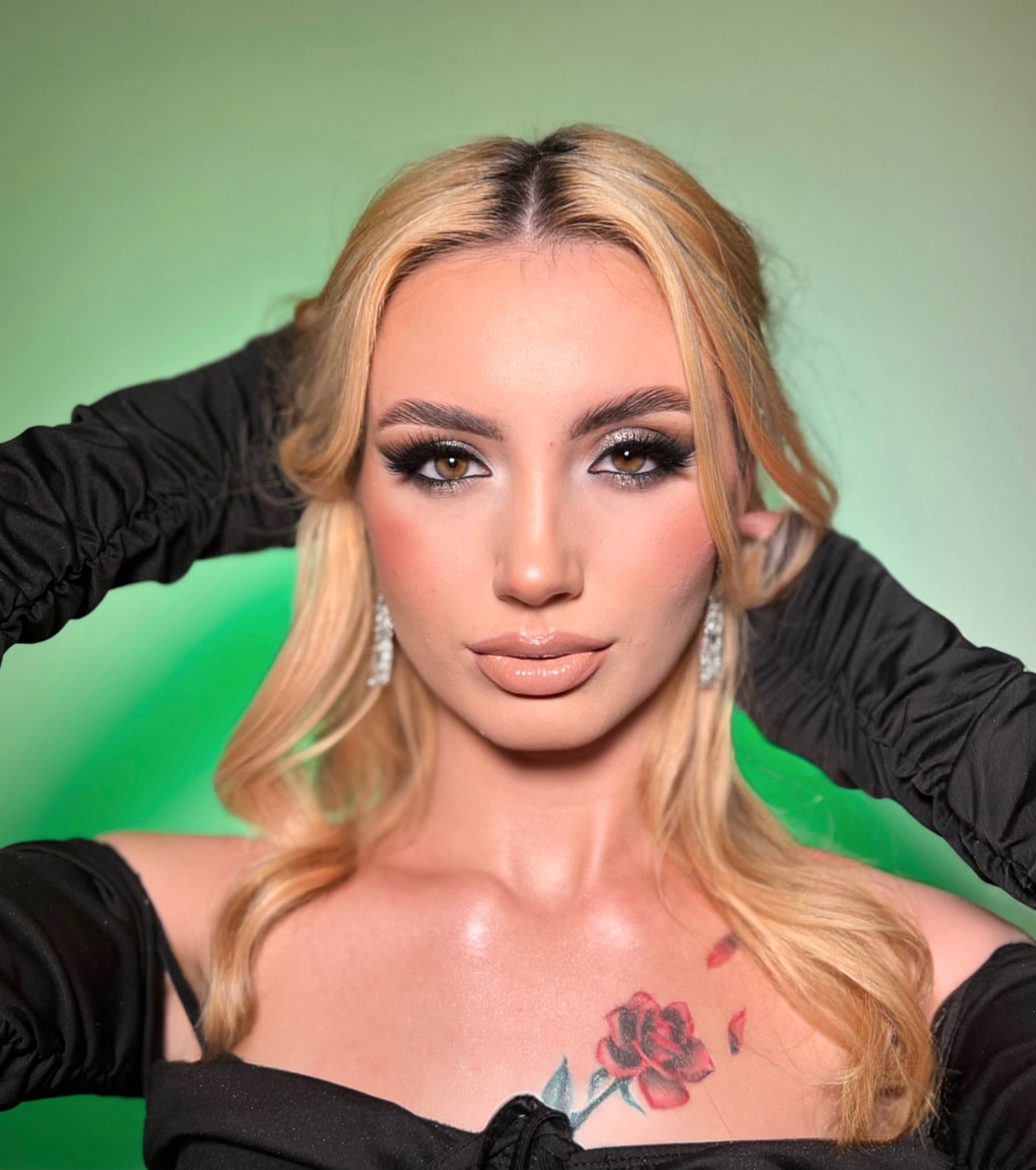 Portret blondwłosej kobiety z wyrazistym makijażem oczu i ust, z tatuażem róży na dekolcie, pozującej na tle zielonego gradientu.
