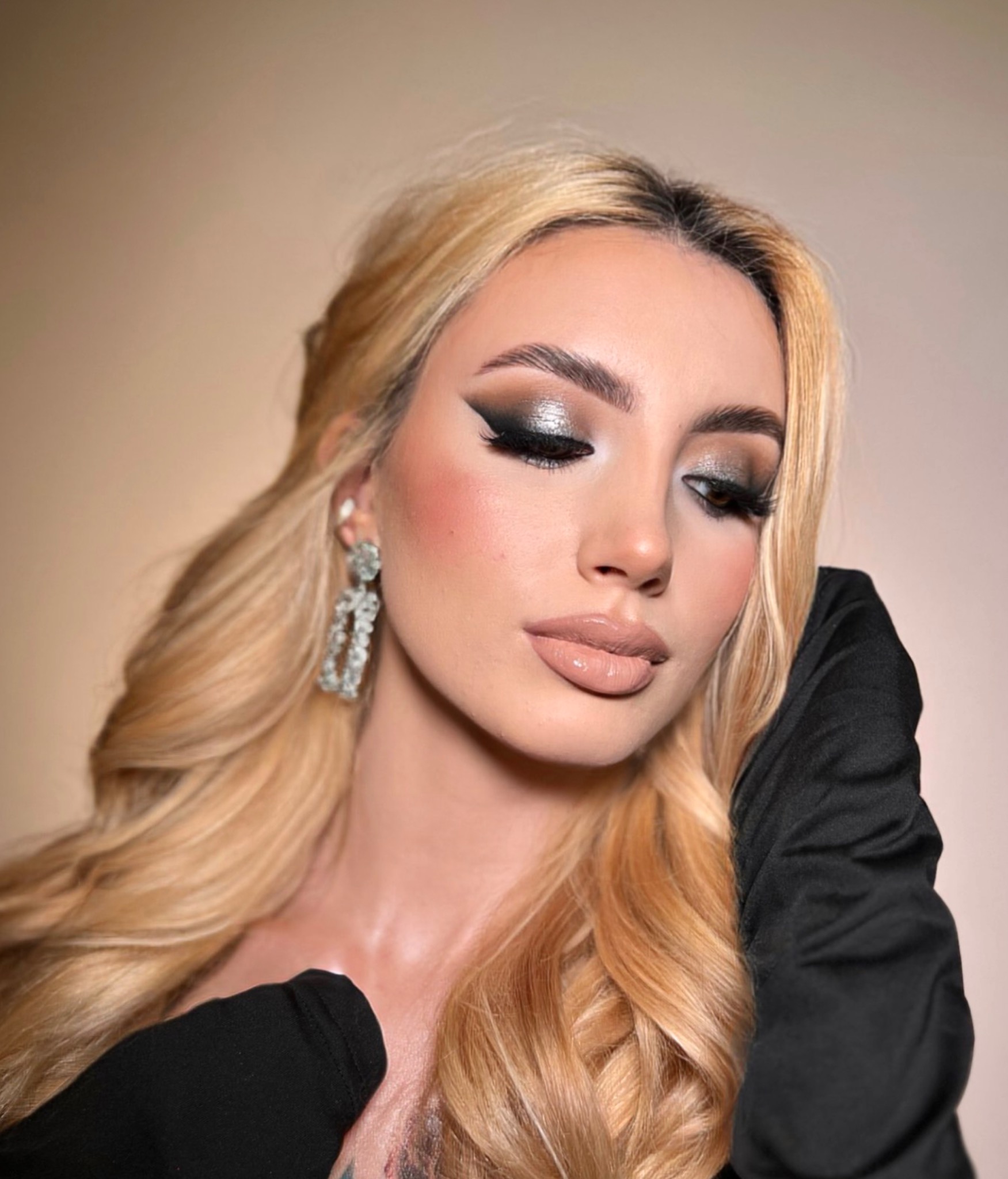 Portret kobiety z platynowymi blond włosami, intensywnym srebrno-czarnym makijażem oczu, błyszczącymi ustami i wiszącymi kolczykami w kształcie kandelabrów.