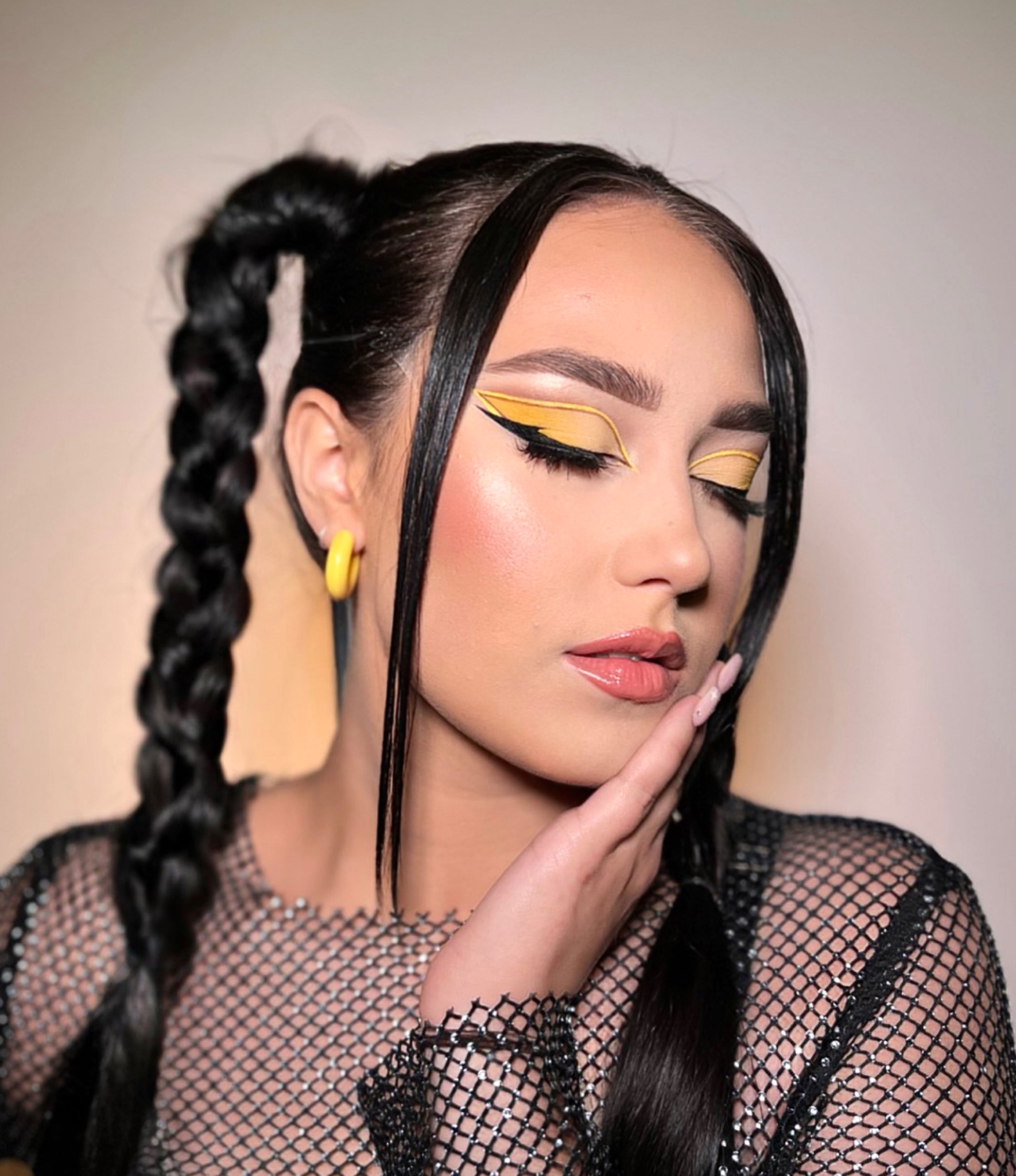 Portret kobiety z zamkniętymi oczami, prezentującej żółto-czarny makijaż oczu i błyszczące usta, z włosami zaplecionymi w dwa warkocze i żółtymi kolczykami kołami.