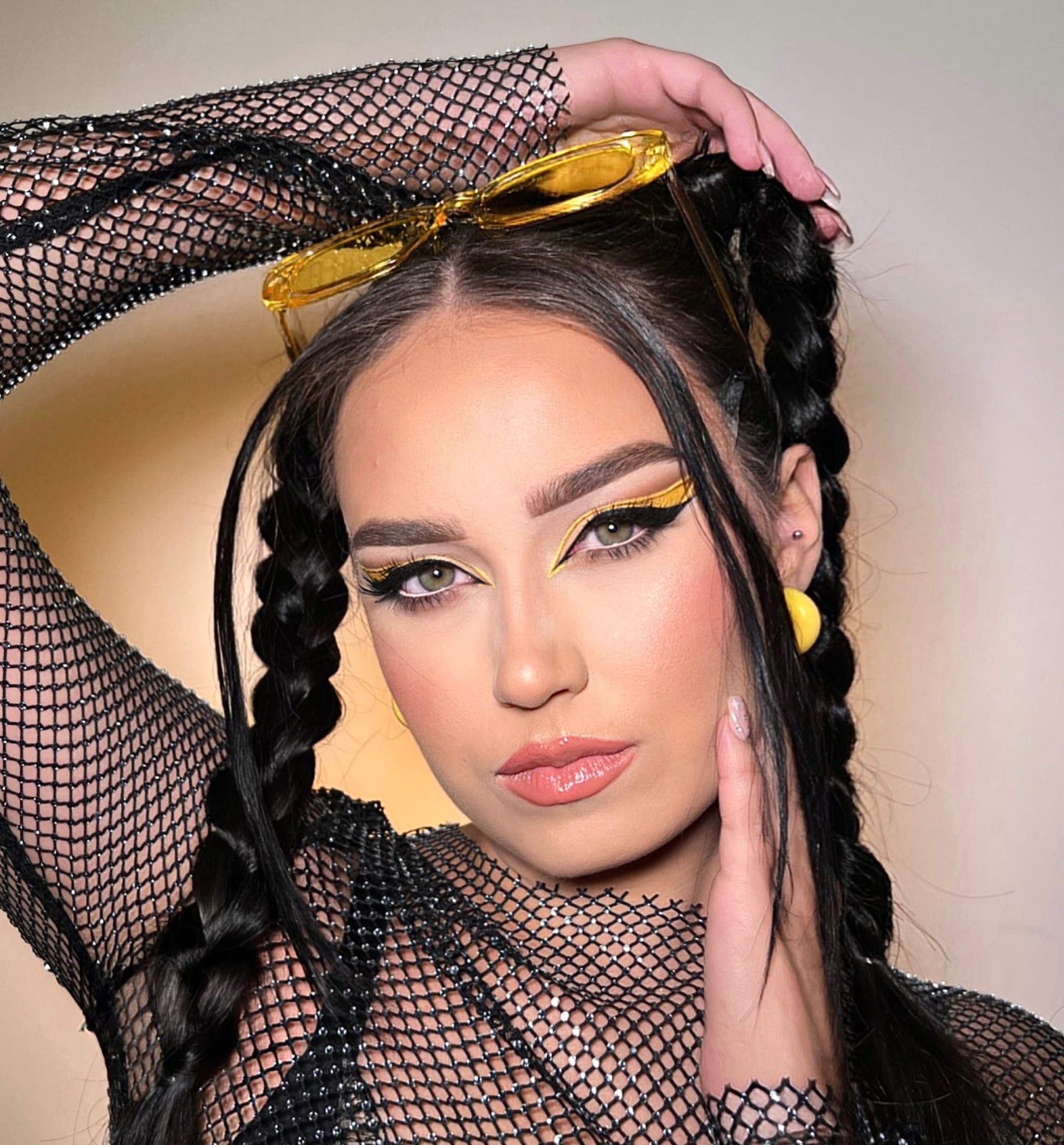 Portret kobiety z wyrazistym, żółto-czarnym makijażem oczu, włosami zaplecionymi w dwa warkocze i okularami na głowie, ubrana w siatkową bluzkę.