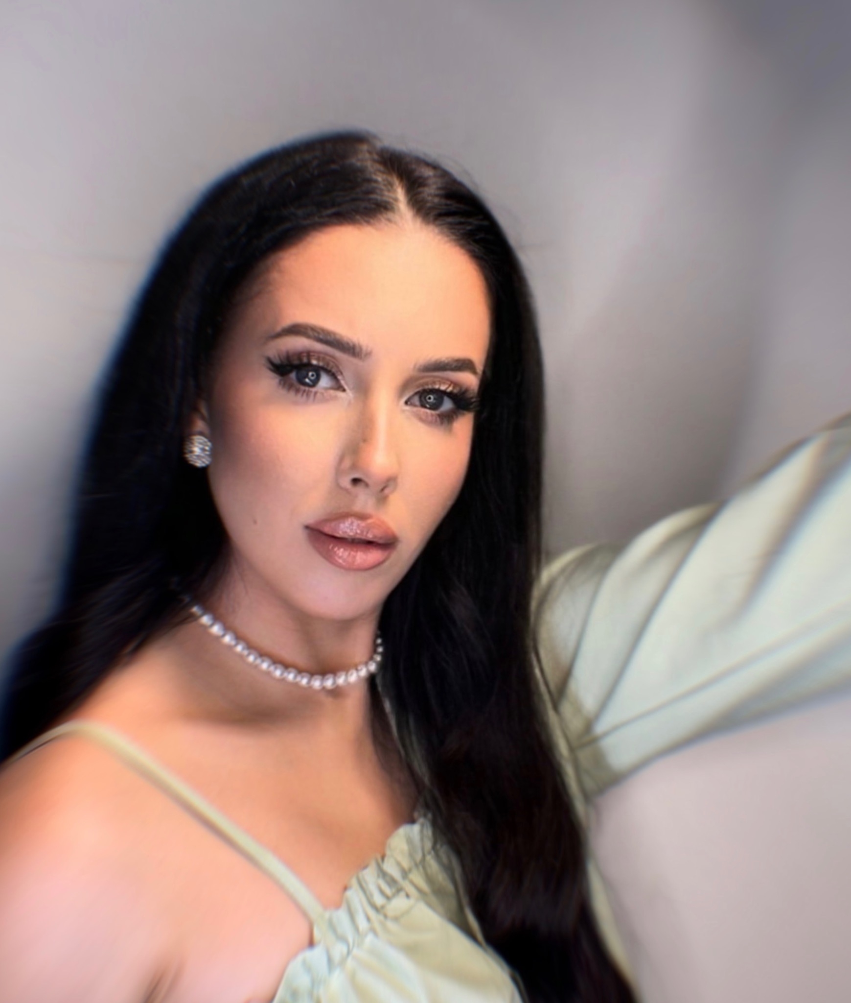 Portret kobiety z wyrazistym makijażem oczu, pełnymi ustami podkreślonymi błyszczykiem i długimi, czarnymi włosami, ubrana w pastelową sukienkę i perłowy naszyjnik.