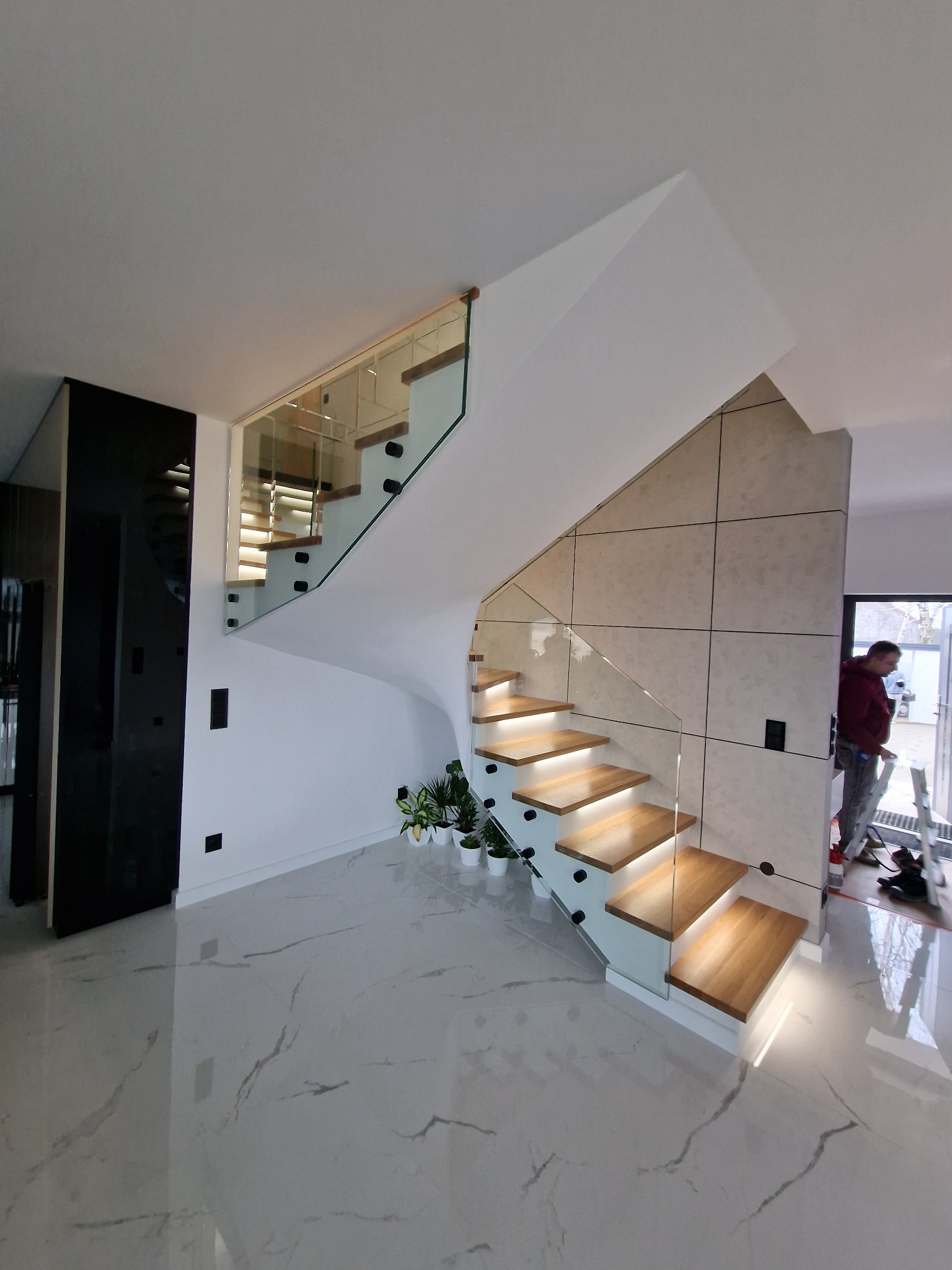 Nowoczesne schody z drewnianymi stopniami podświetlonymi od spodu i szklaną balustradą, widoczne rośliny doniczkowe i pracownik na drabinie.