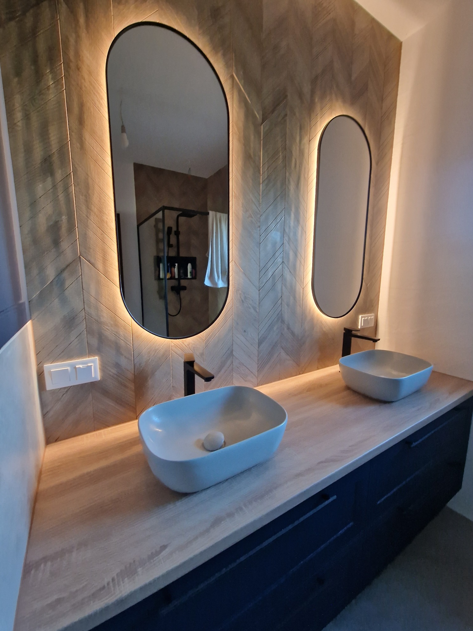 Nowoczesna łazienka z dwiema umywalkami nablatowymi, podświetlanymi lustrami w czarnych ramach i ścianą wyłożoną płytkami imitującymi drewno ułożonymi w jodełkę.