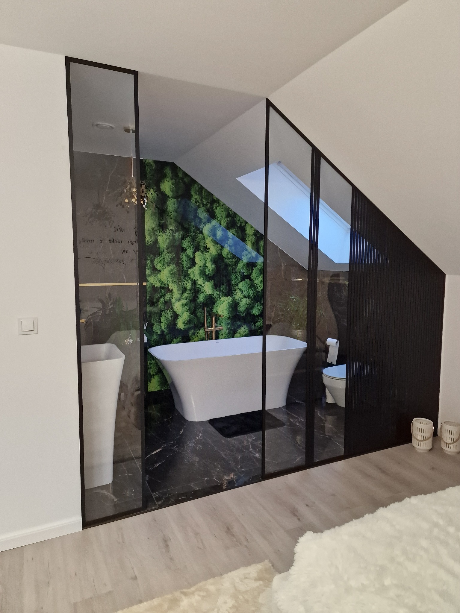 Nowoczesna łazienka na poddaszu oddzielona szklaną ścianką działową z czarnymi ramami, z wolnostojącą wanną, umywalką na postumencie i akcentem zielonej ściany z mchu.